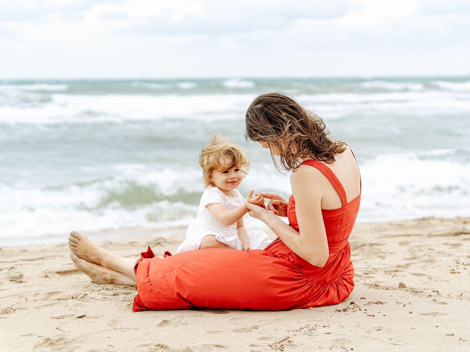 Mother in red dress sitting on beach sand interacting with her toddler near ocean waves.