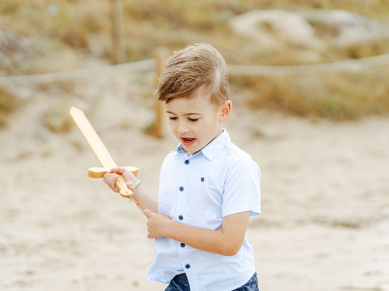 Young boy in blue shirt examining wooden toy airplane on sandy beach.