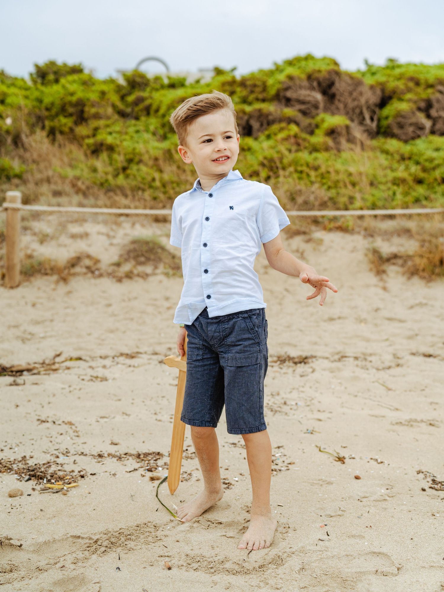 Young boy in light blue button-up shirt and navy shorts holding wooden toy on sandy beach.