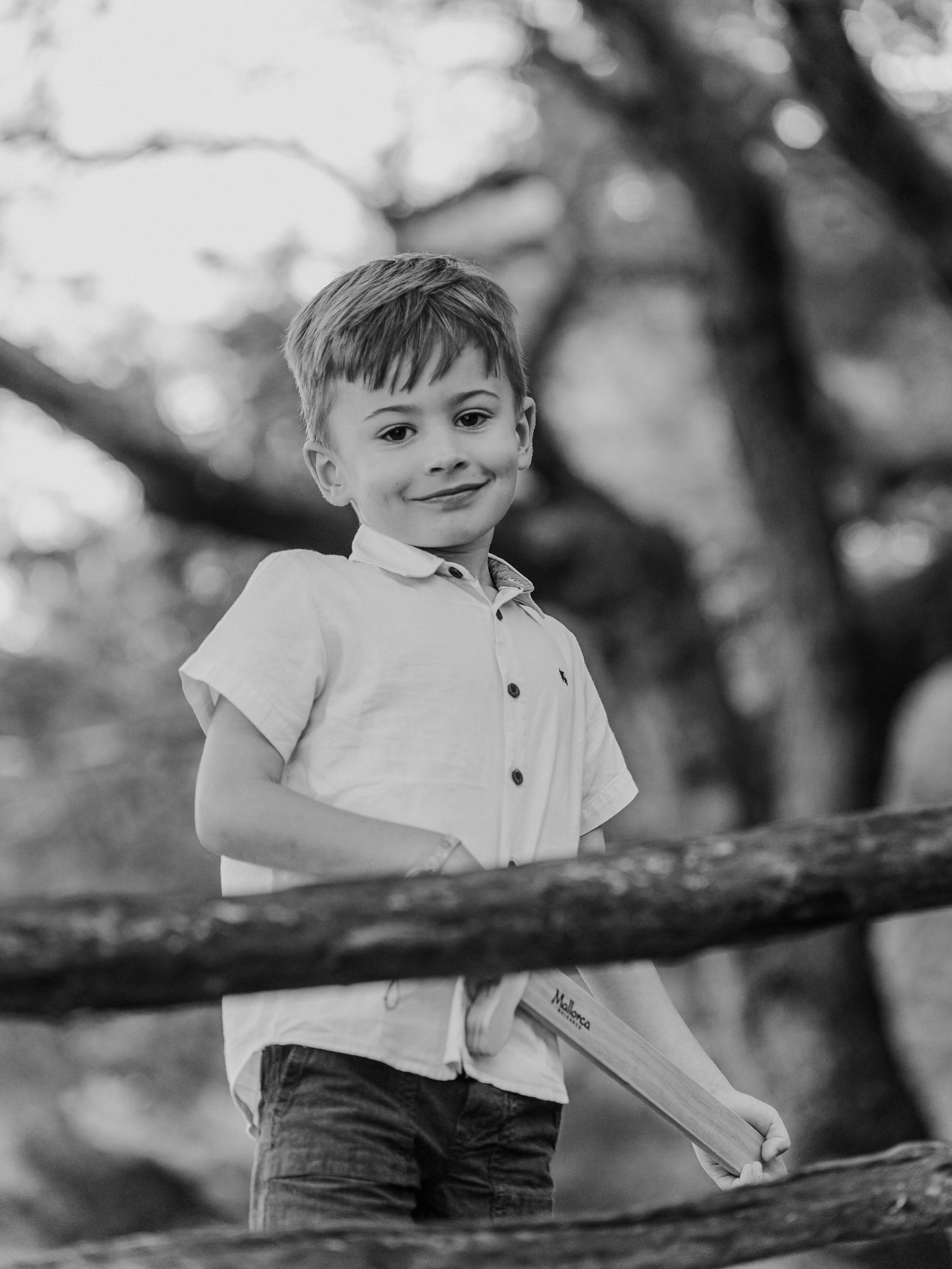 Black and white portrait of a smiling boy holding a wooden toy while leaning on a rustic fence.