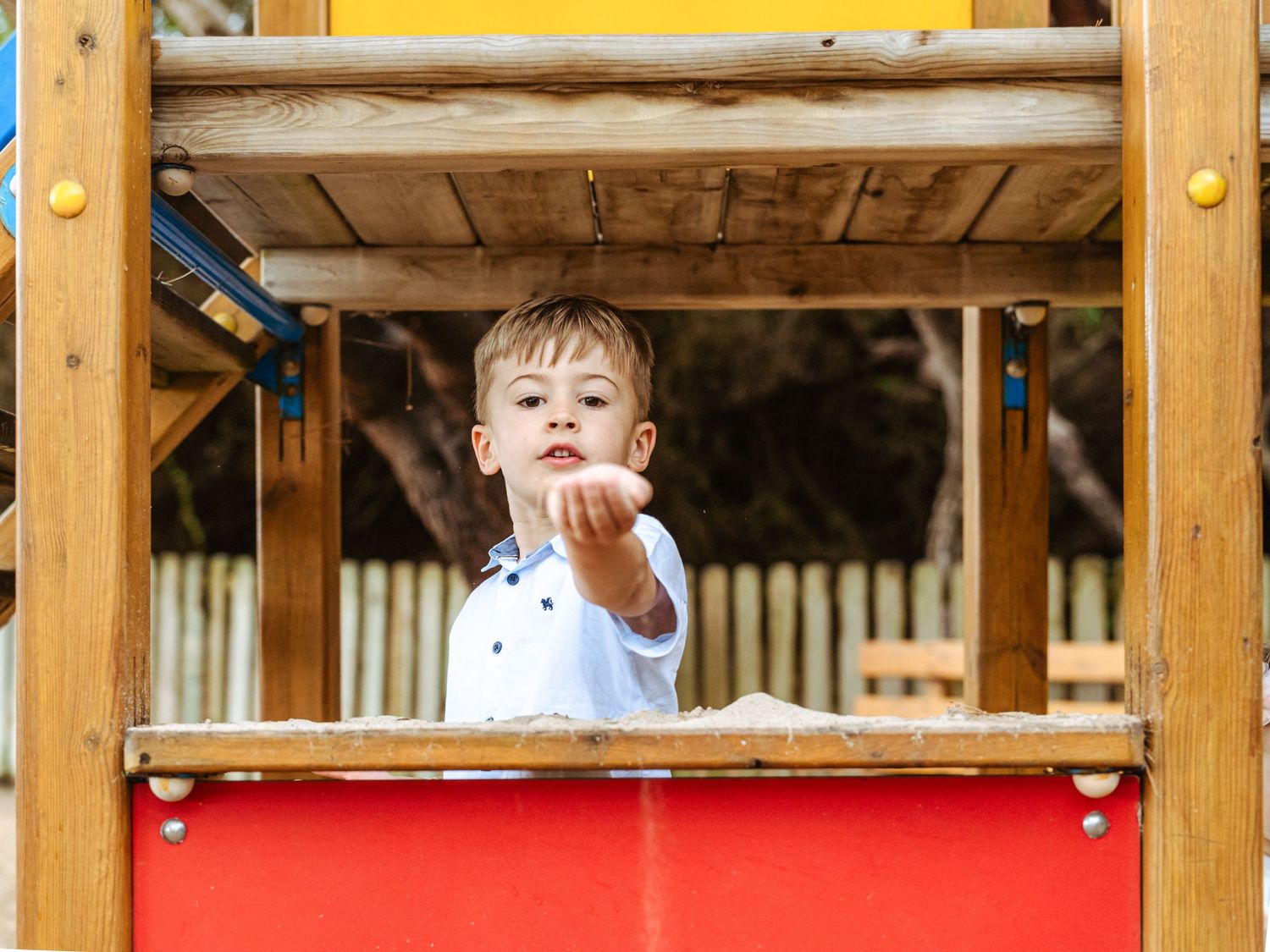 Young boy in white shirt pointing from wooden playground structure, colorful red and yellow panels visible.