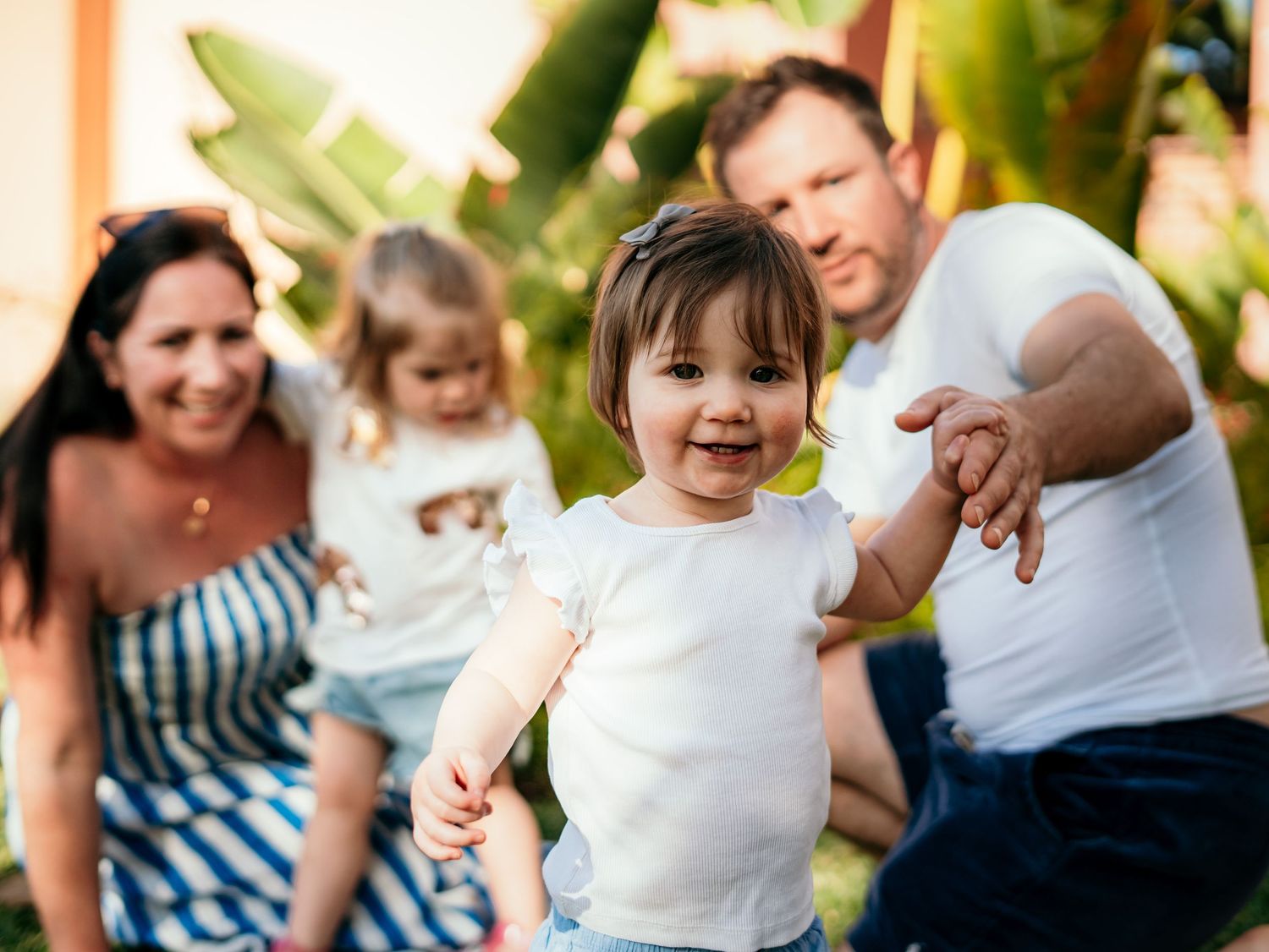 Joyful toddler girl in white top running toward camera with family smiling behind her.