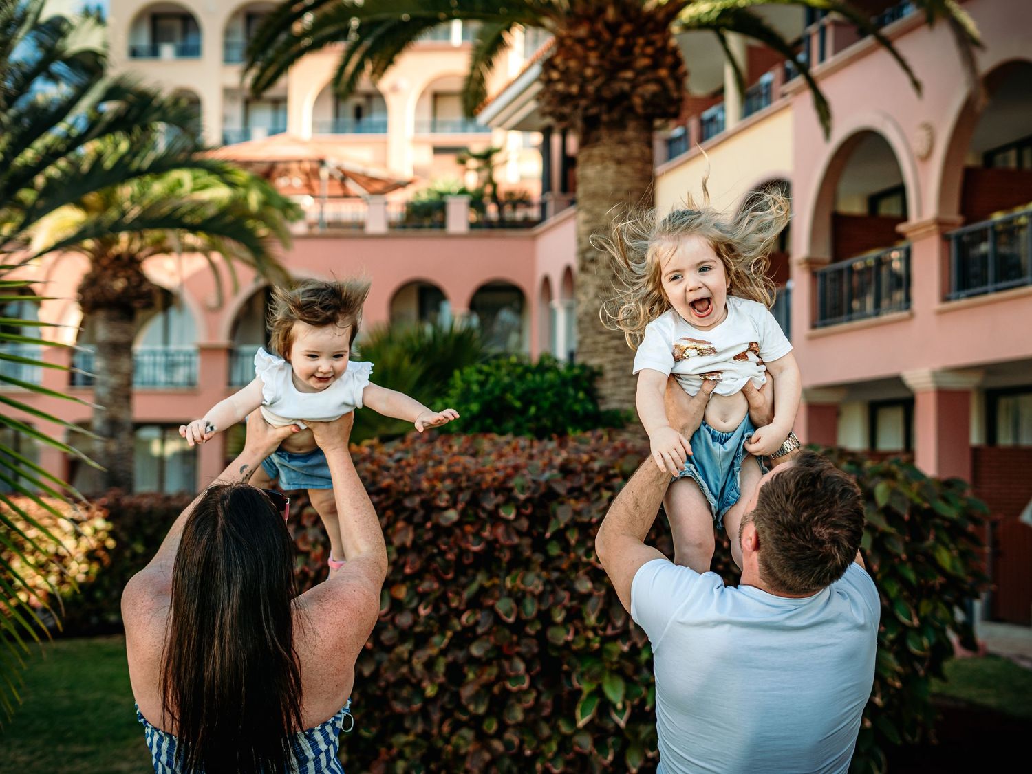 Parents joyfully lift two laughing young children in the air outside a tropical resort.
