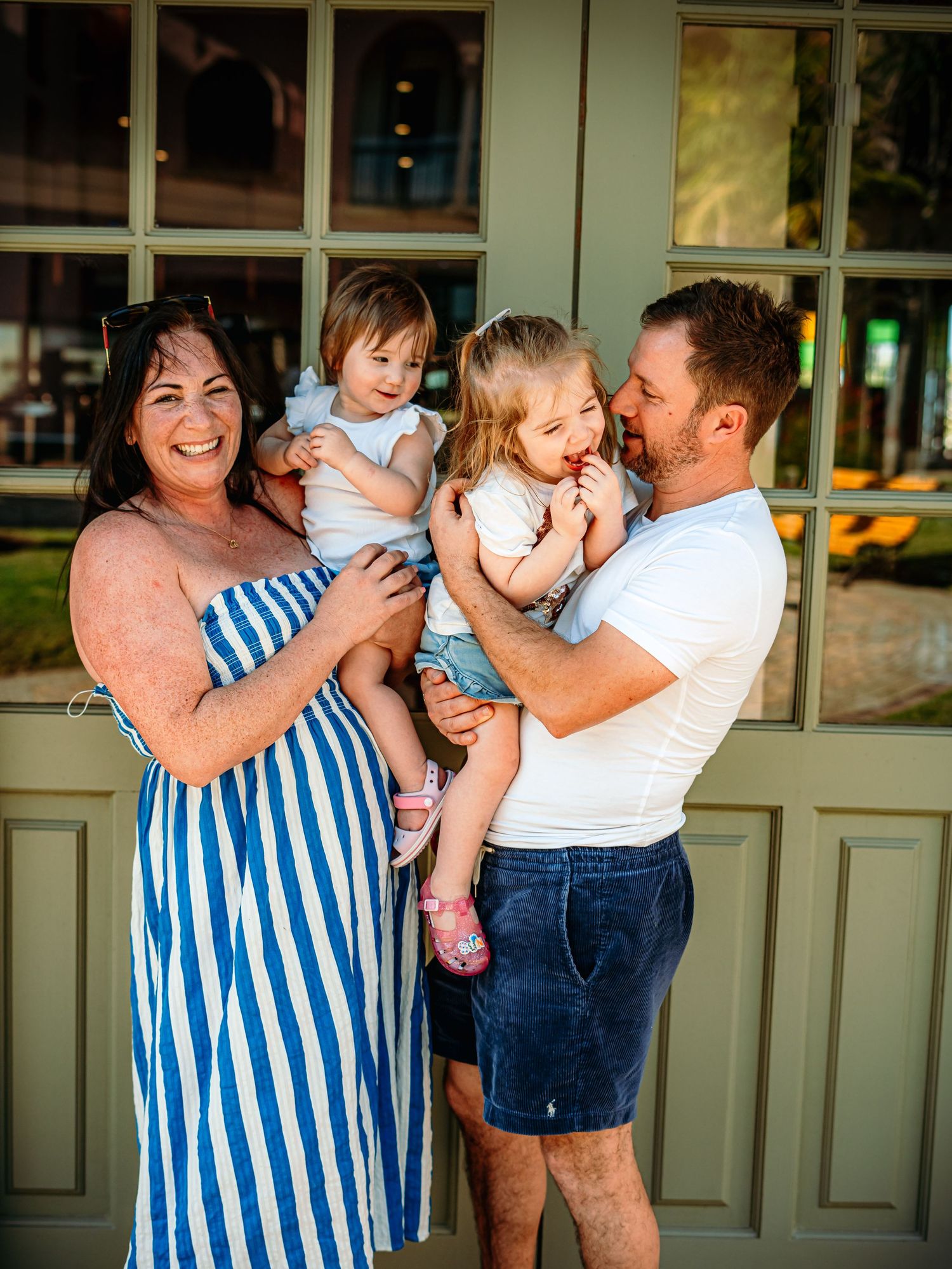 Happy family of four with parents holding two young daughters smiling in front of green French doors.