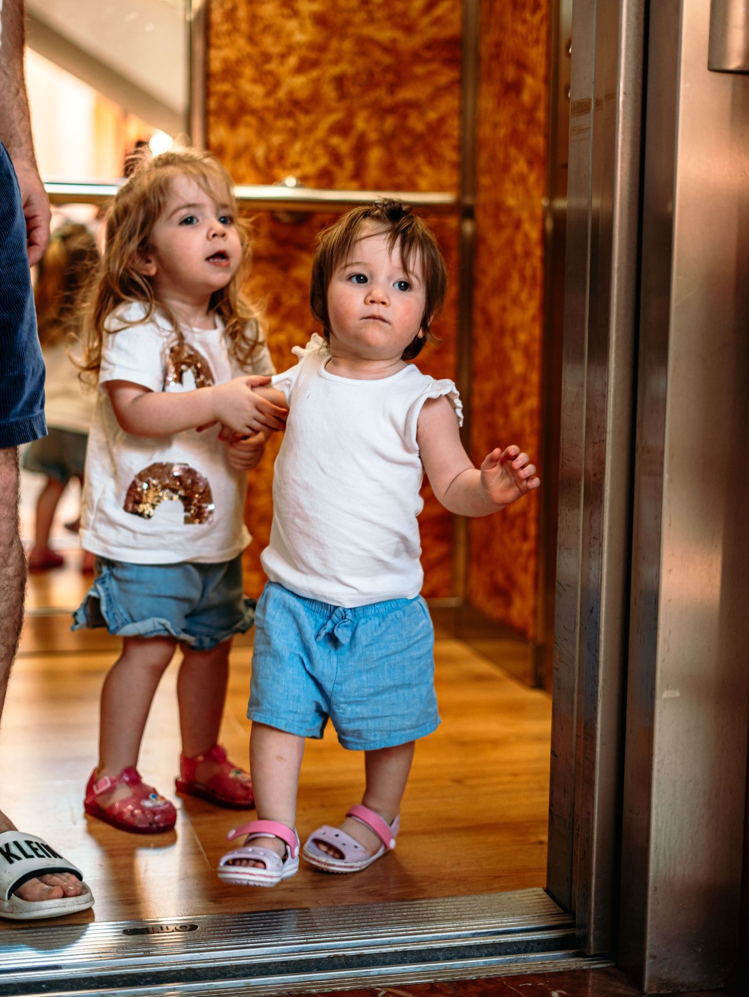 Two toddlers in matching white tops and blue shorts standing at elevator doors.