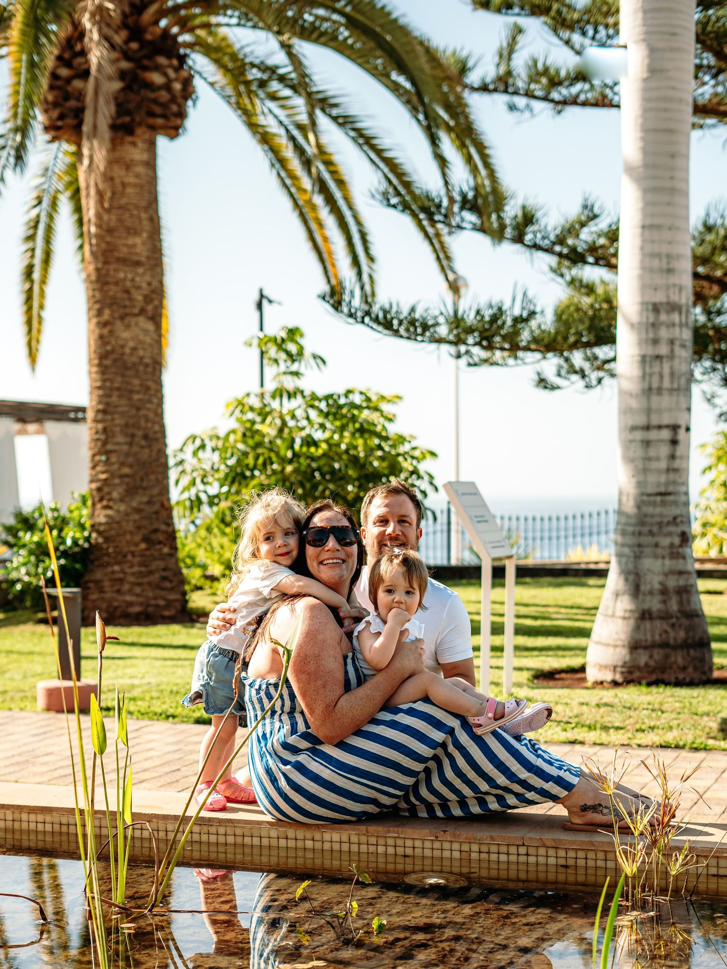 Joyful family of four sitting together by a garden pond with tall palm trees behind them.