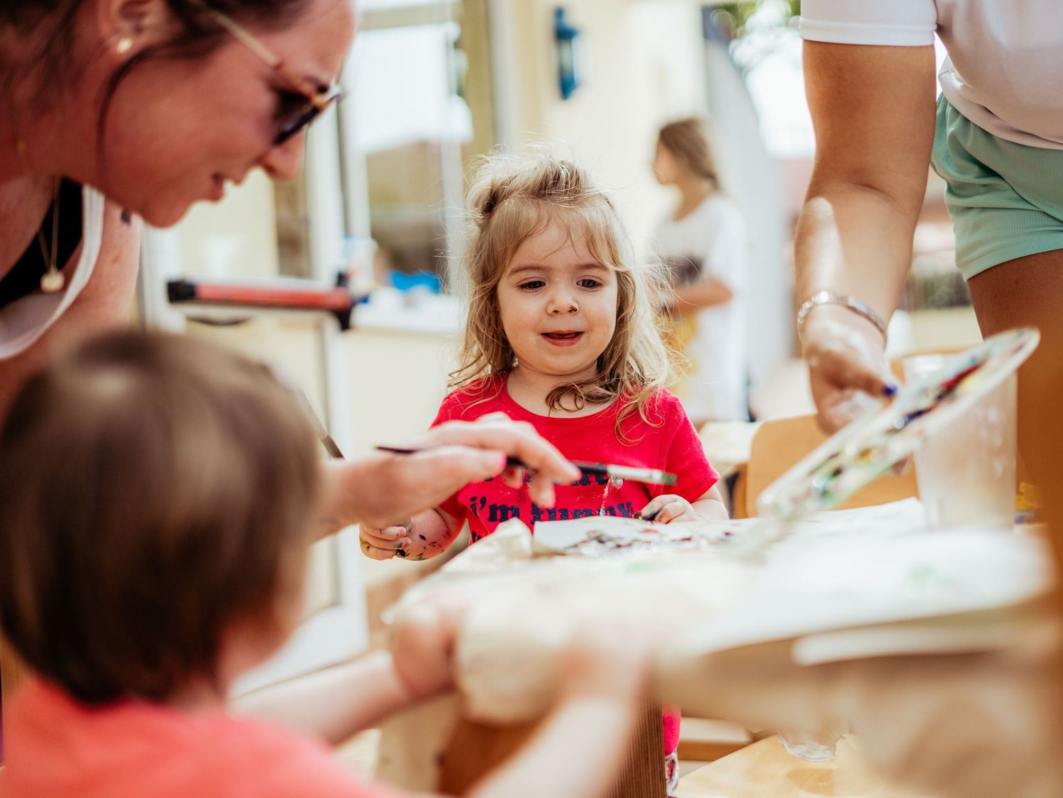A toddler girl in red smiles while two adults assist her with a creative painting activity.