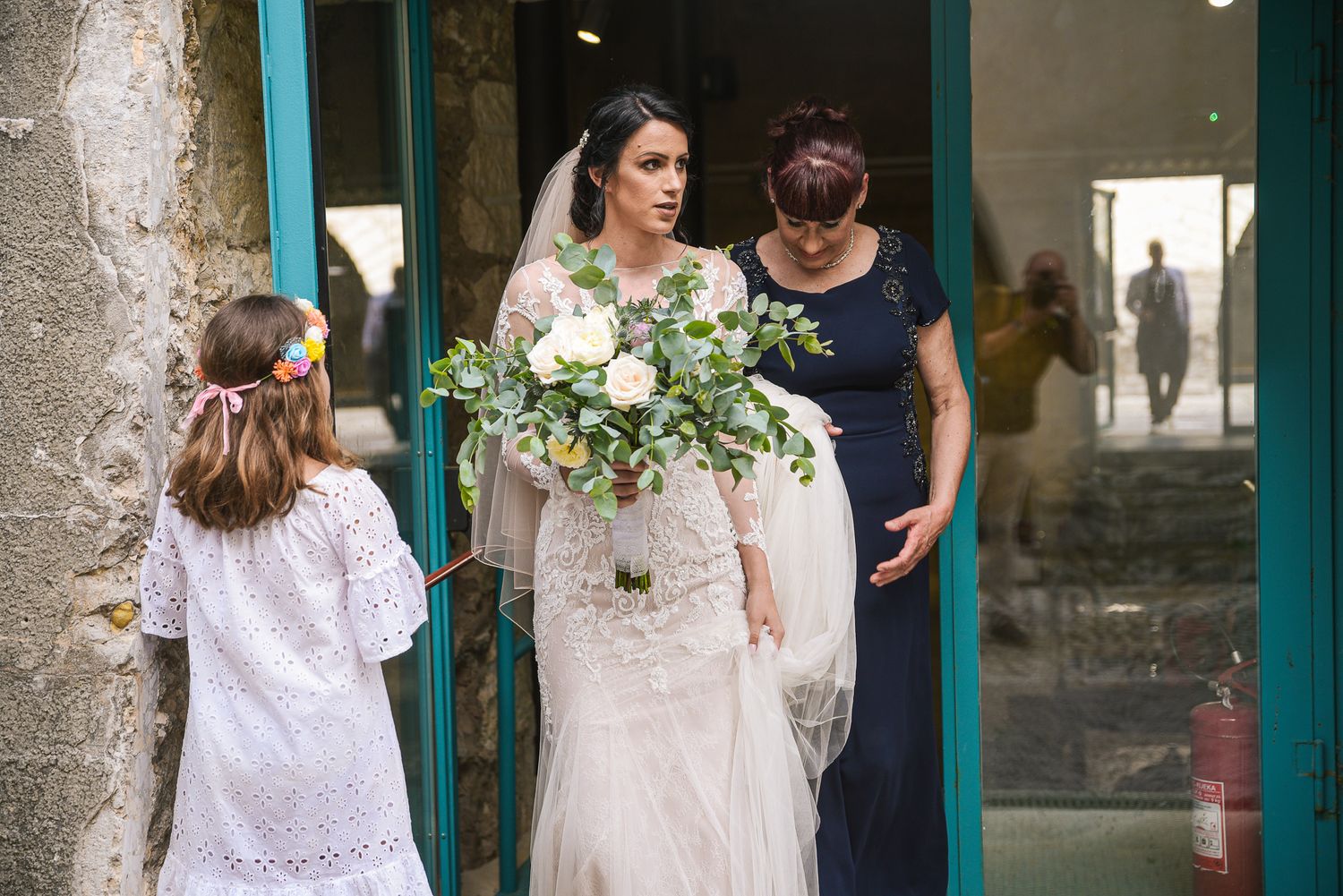 Bride in lace gown holding white bouquet exits doorway with bridesmaid and flower girl.