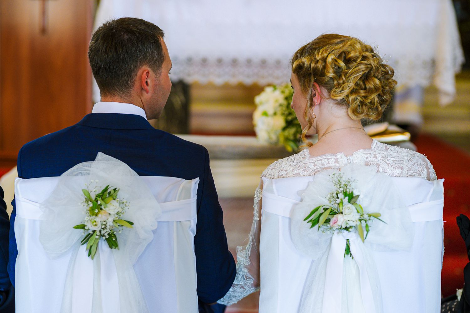 Back view of bride and groom seated in white ribbon-adorned chairs during church ceremony.