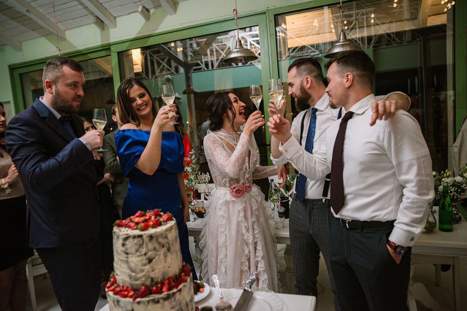 Wedding guests toast with champagne beside a strawberry-decorated two-tier naked cake.