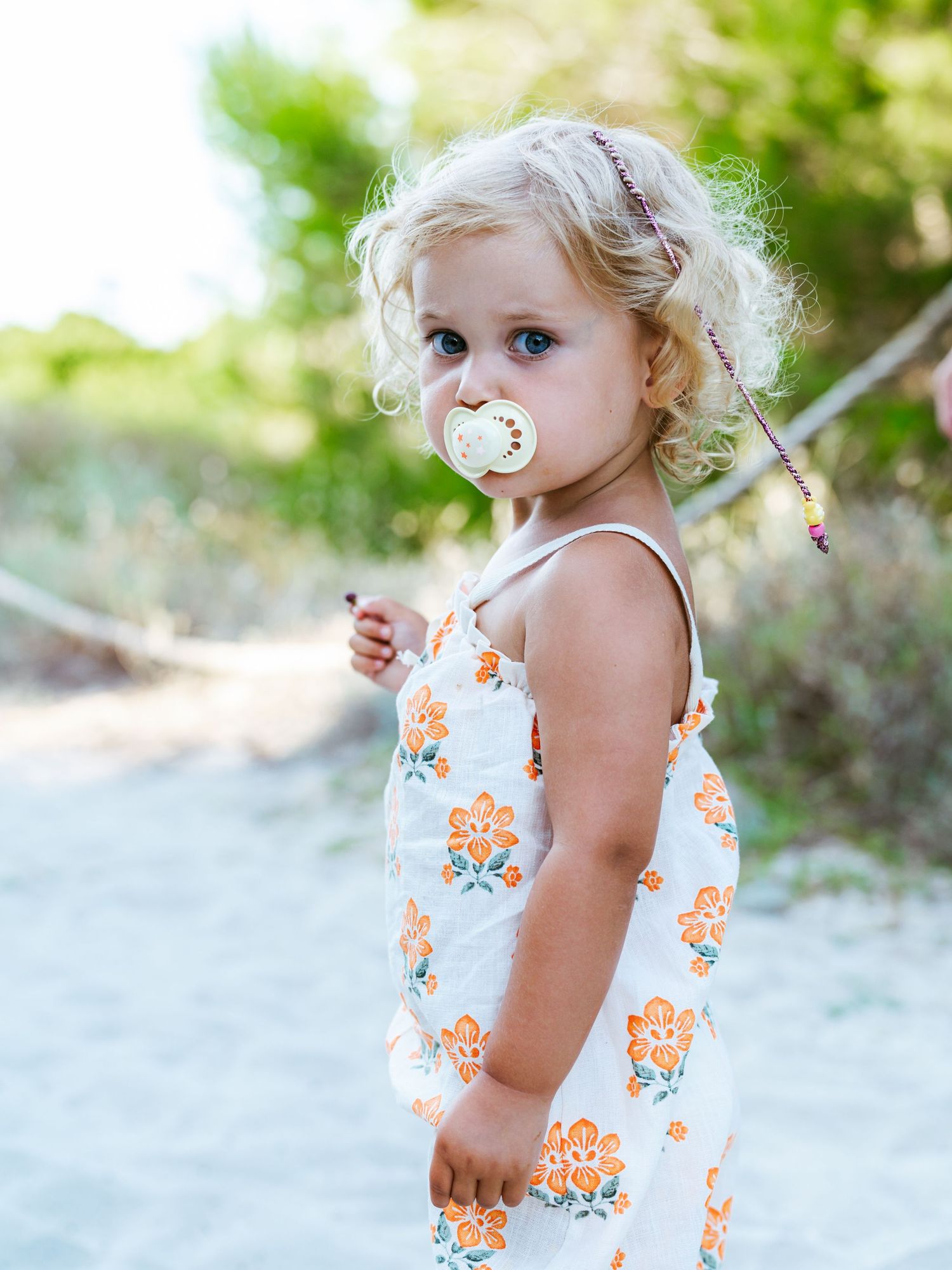 Blonde toddler girl with pacifier wearing floral sundress standing on sandy beach path.