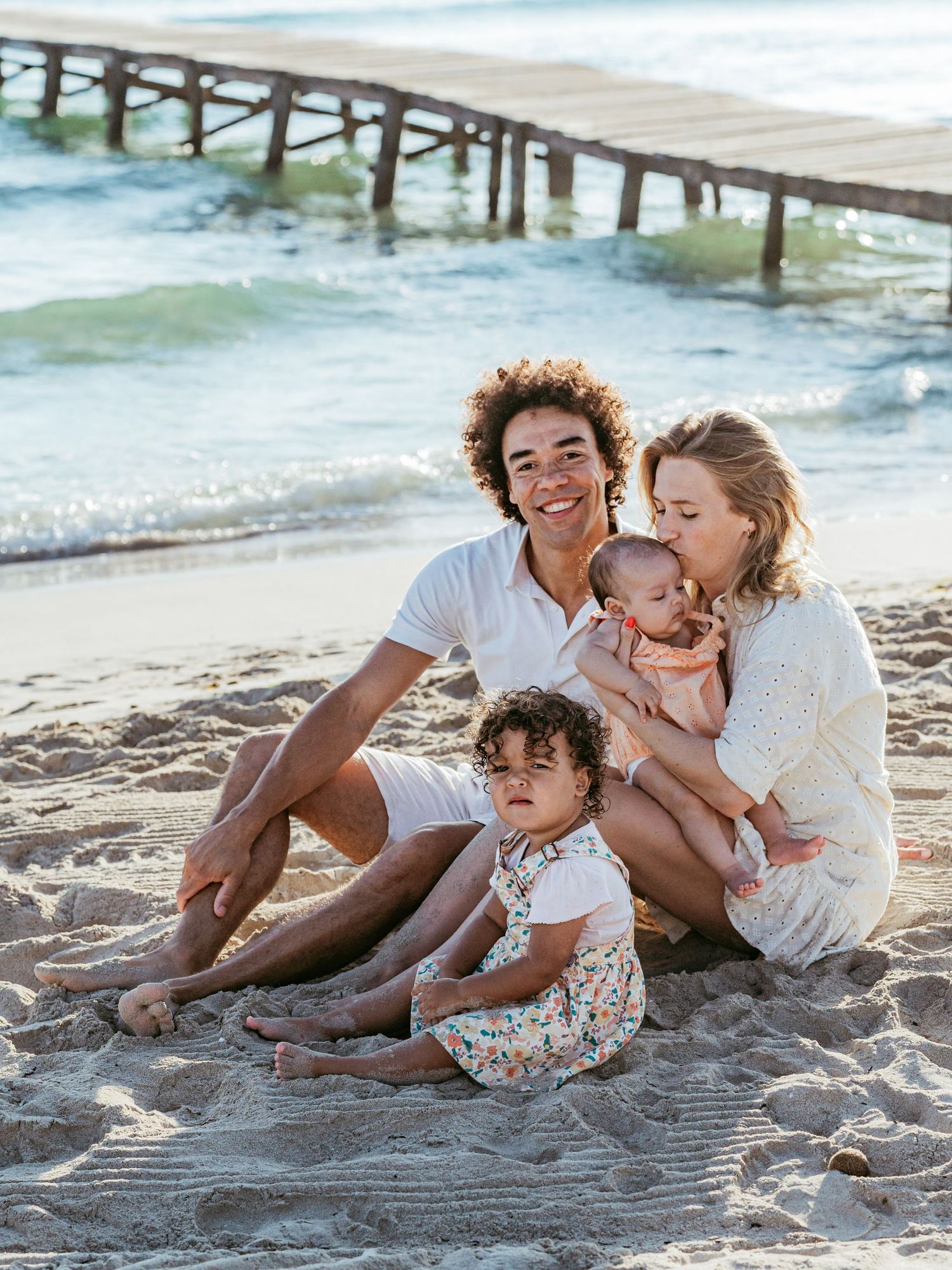 A happy family of four sits on a sandy beach, with parents holding two young children near a wooden pier.