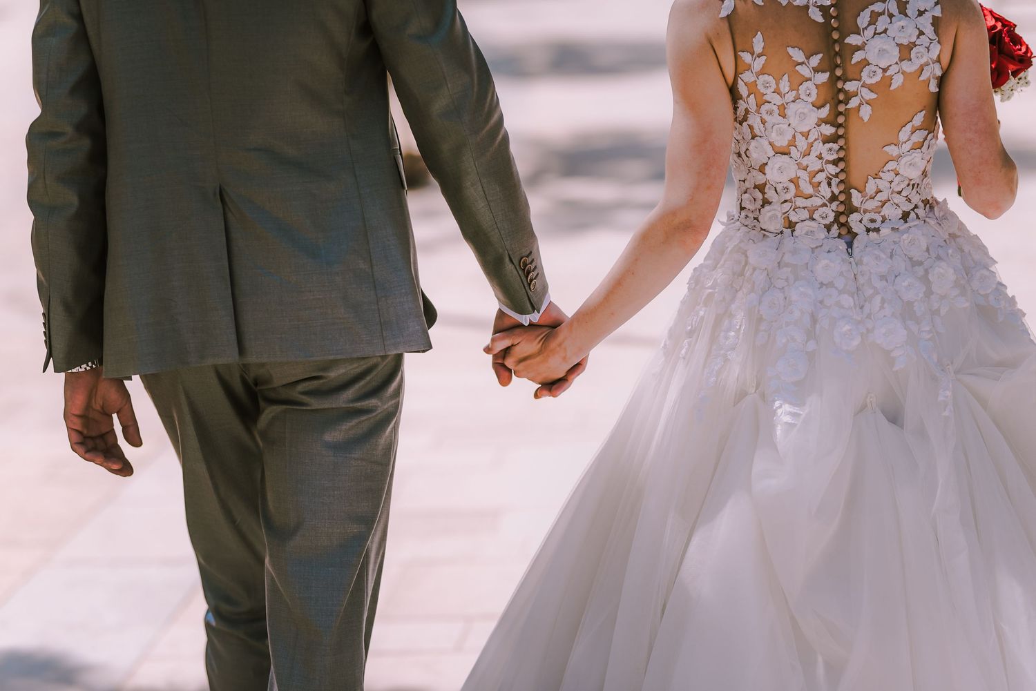 Bride and groom hold hands walking, showcasing lace backless wedding dress and gray suit.