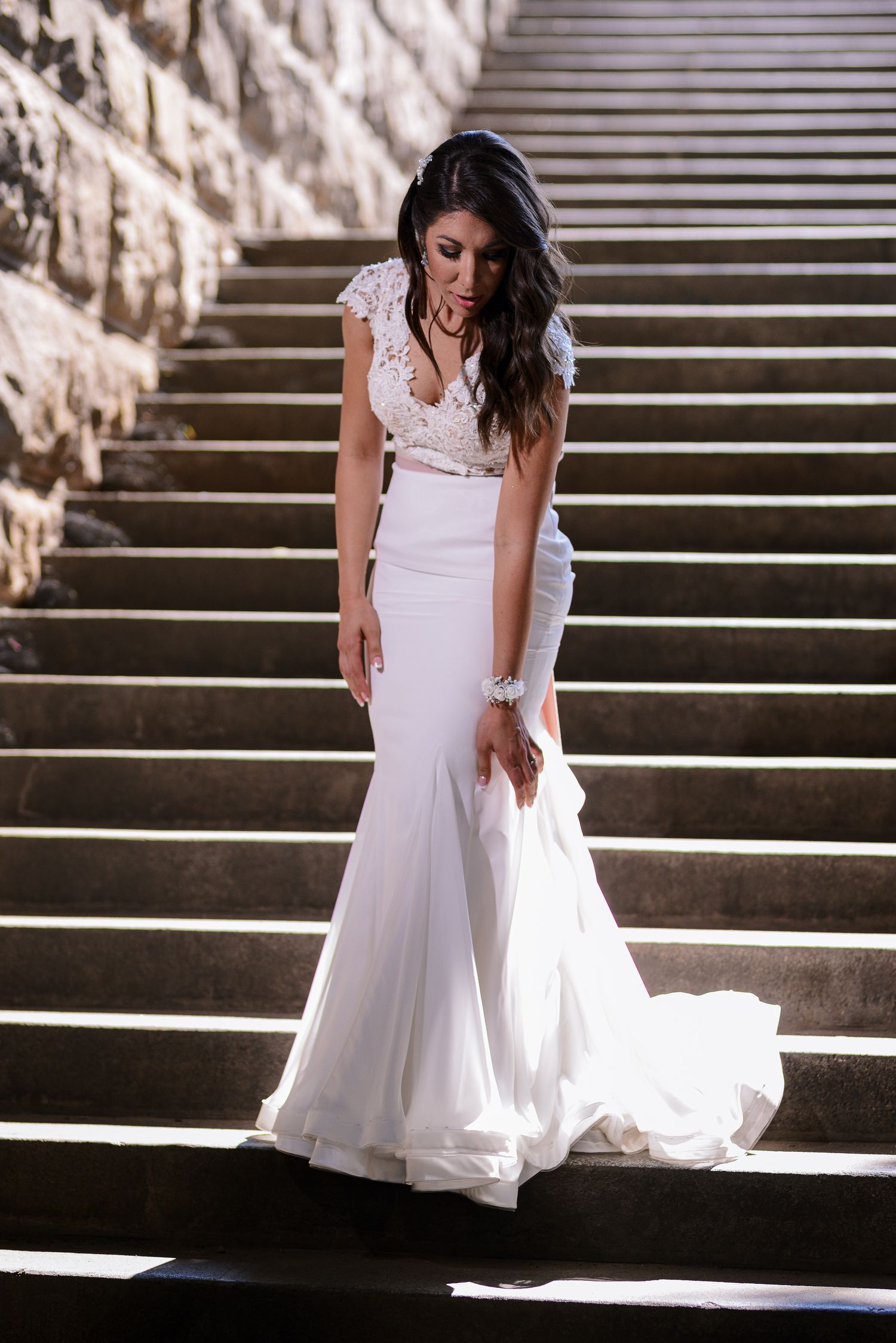 Elegant bride in a fitted lace mermaid wedding gown poses gracefully on sunlit stone steps.