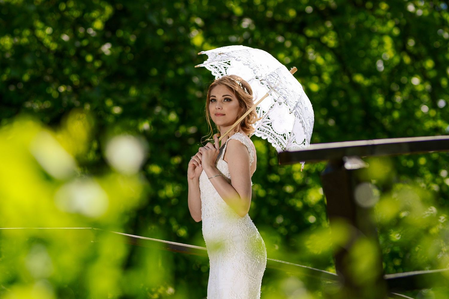 Elegant bride in a white dress holds a decorative lace parasol umbrella while standing in a lush green garden.