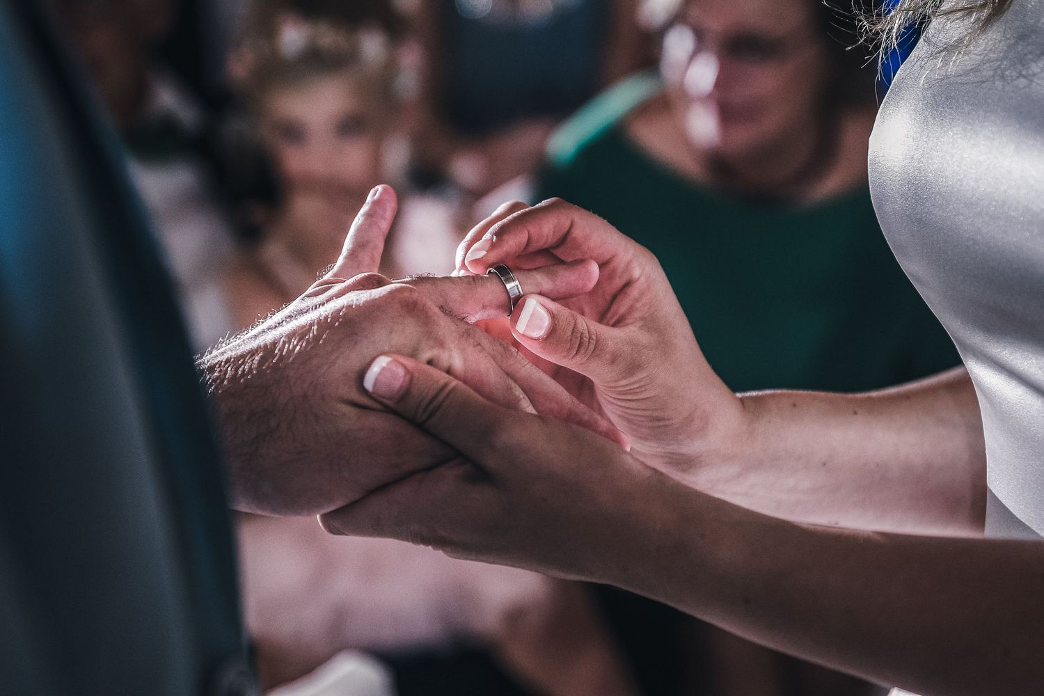 Close-up of wedding ring exchange ceremony with guests visible in the background.