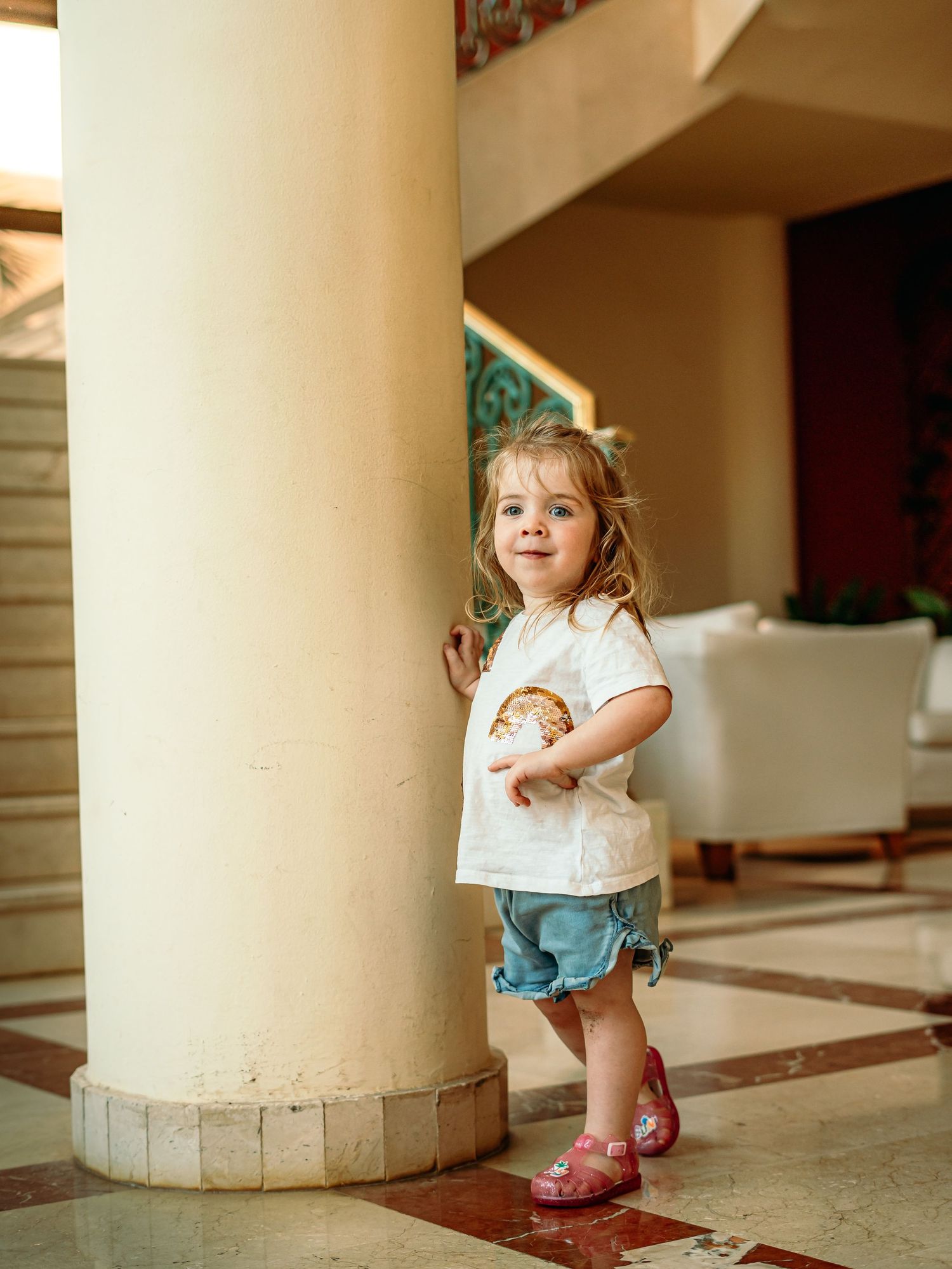 A smiling toddler girl in a white graphic tee and denim shorts leans against a large column indoors.
