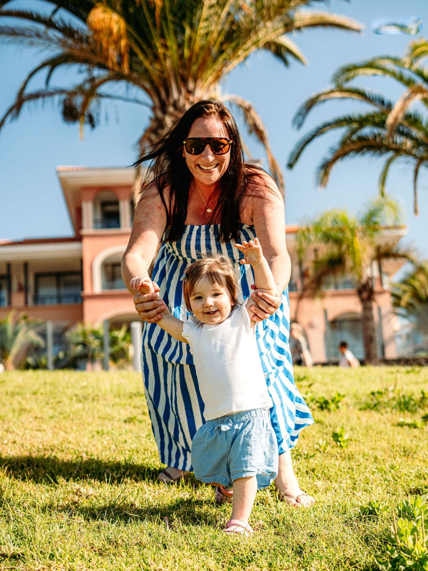 A laughing woman in a blue striped dress holds a toddlers hands as they walk on sunny grass.