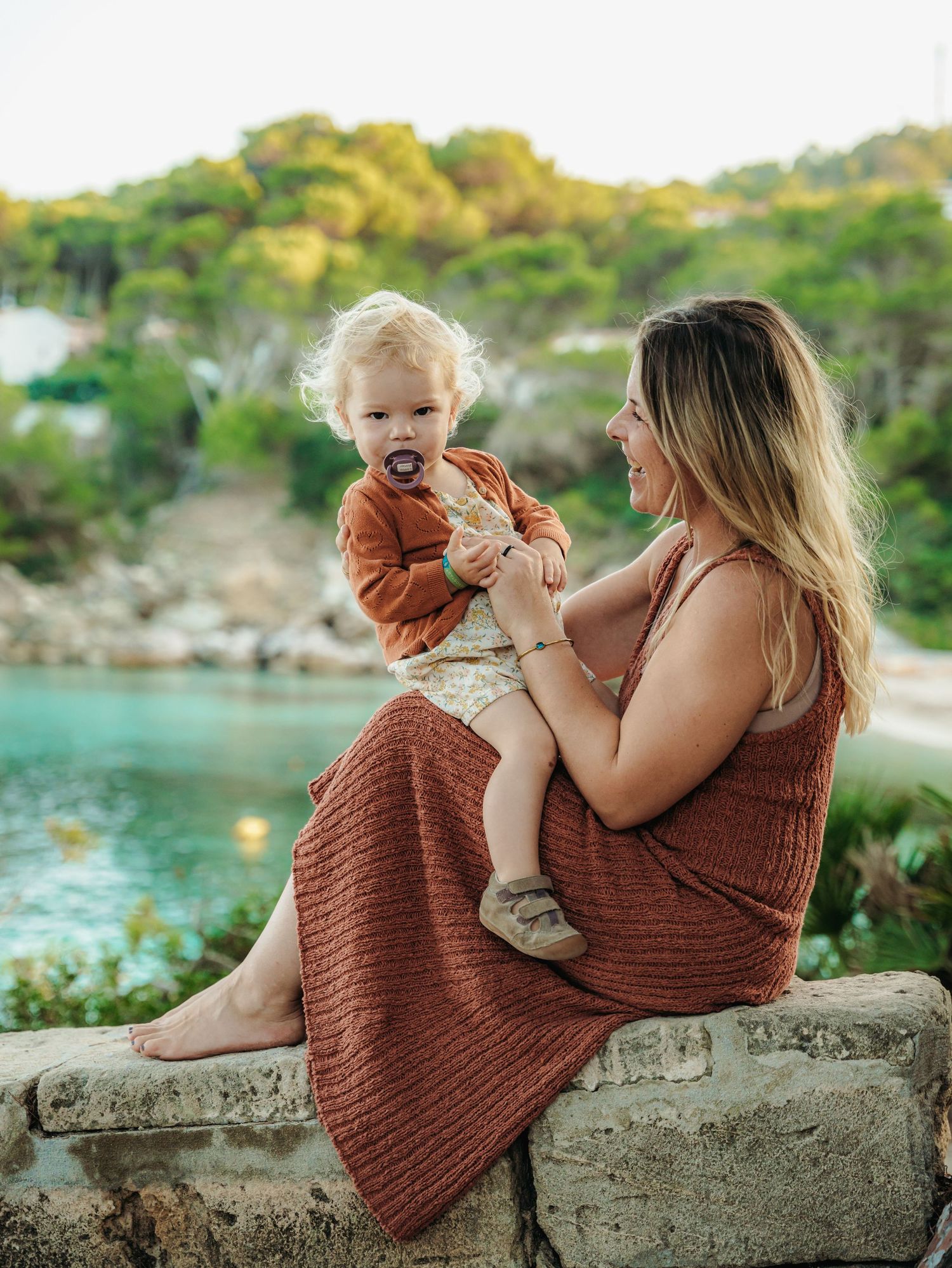 Mother in rust knit maxi dress sitting on stone wall holding toddler near scenic cove.