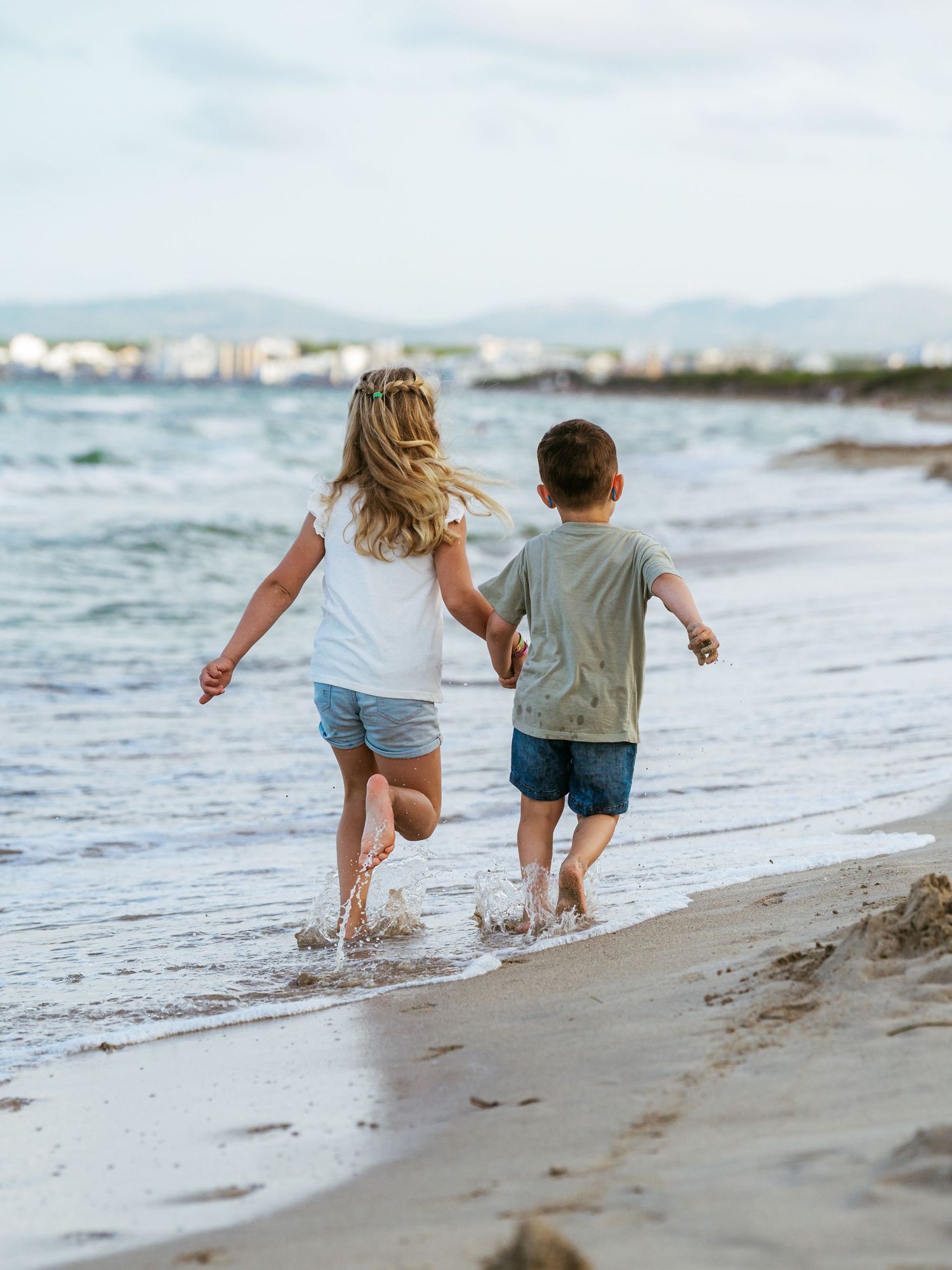Two young children holding hands running along shallow ocean waves on a sandy beach.