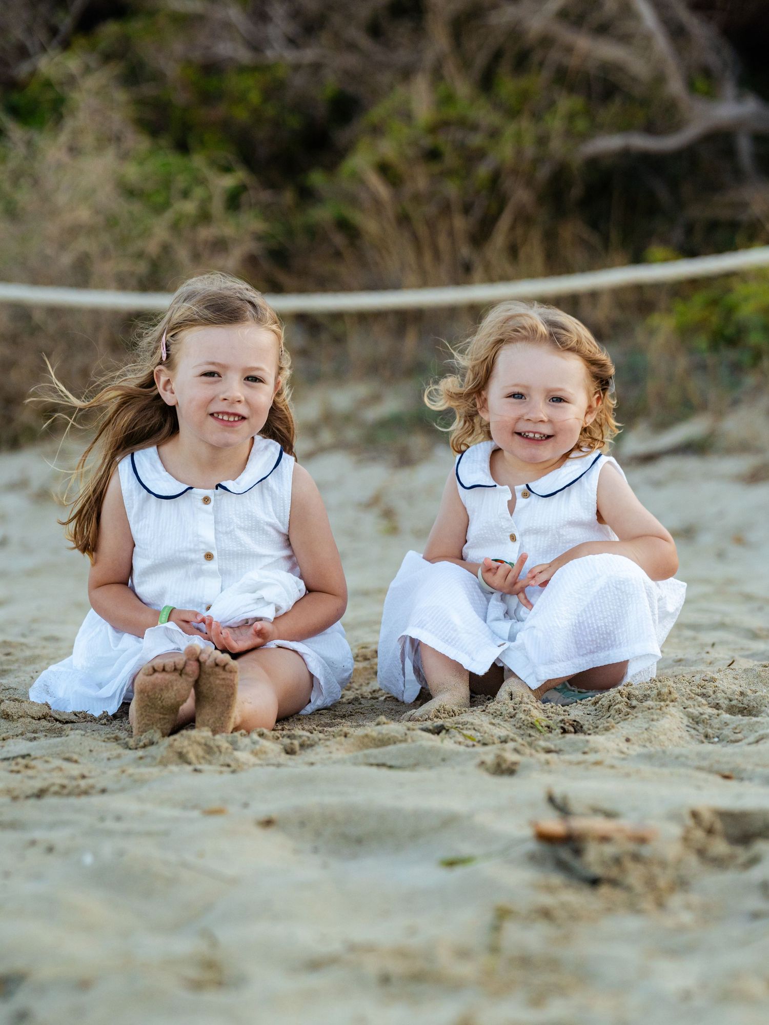 Two young sisters in matching white dresses sitting together smiling on sandy beach.