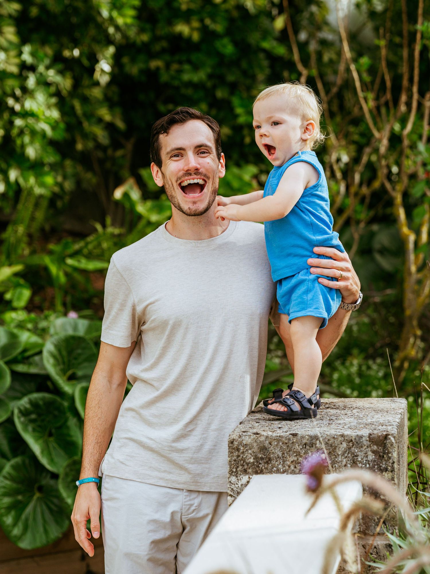 Laughing father lifting his excited toddler in a blue outfit in a lush garden setting.