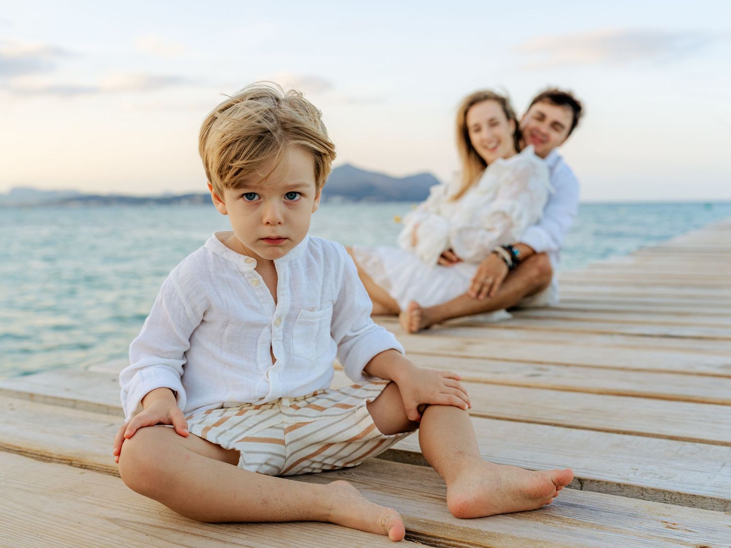 Young boy sits cross-legged on a wooden pier while his parents relax together in the background by the sea.