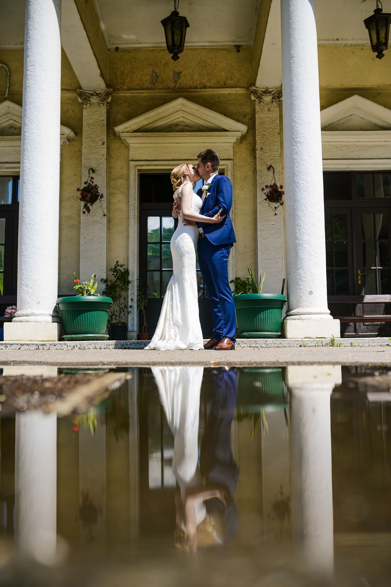 Bride and groom share a romantic kiss reflected in a pool of water before a grand columned estate.