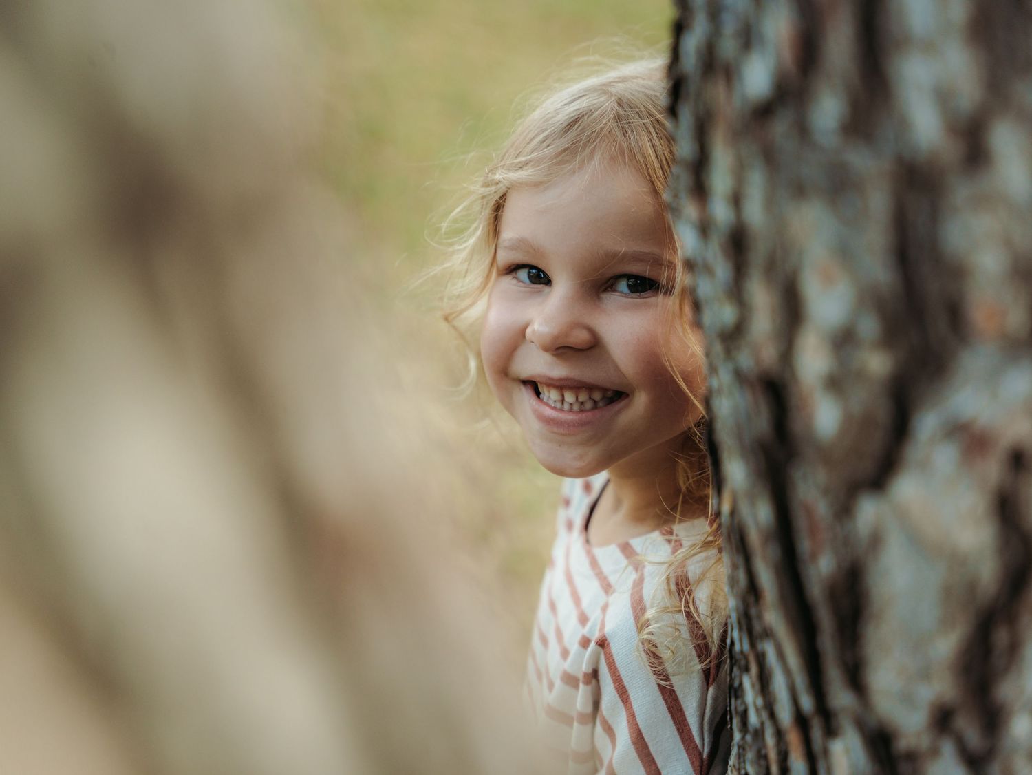 Smiling blonde girl peeking from behind a tree trunk in a forest setting.