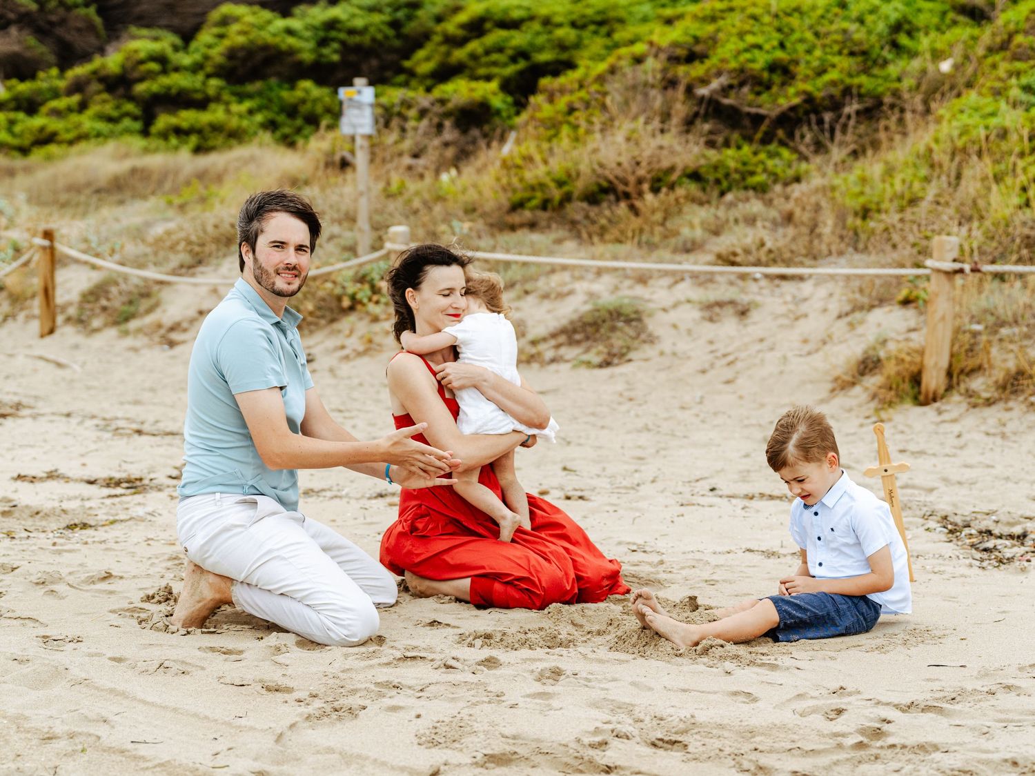 Family of three playing in sand on beach, parents in blue and red outfits with toddler son.