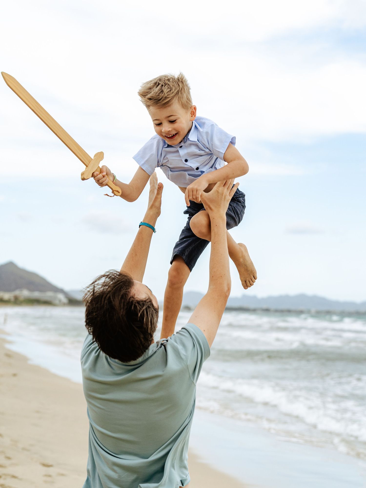 Father lifting smiling boy holding wooden sword high above his head on a sandy beach.