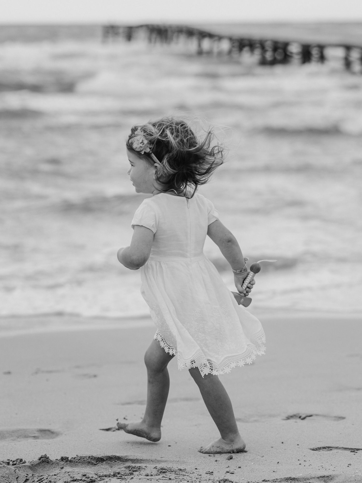 Black and white photo of young girl in white dress running barefoot along beach near ocean waves.
