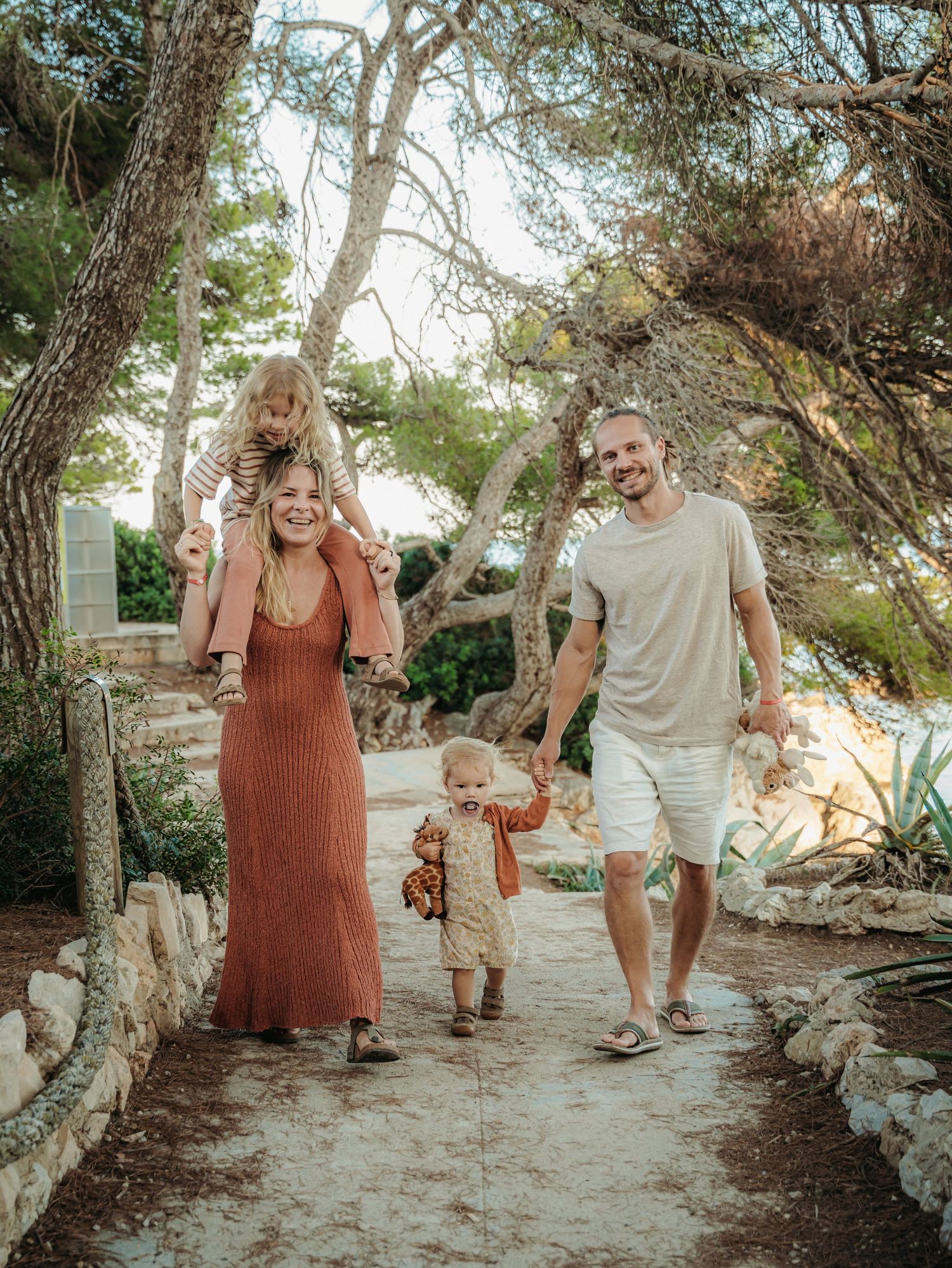 Happy family walks hand-in-hand on sunlit stone path through pine trees, mother with child on shoulders.
