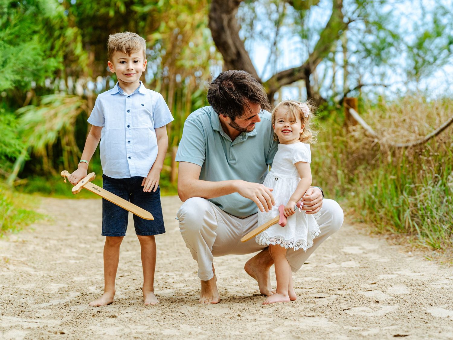 Smiling father kneeling on sandy path between his young son with wooden sword and laughing toddler daughter.