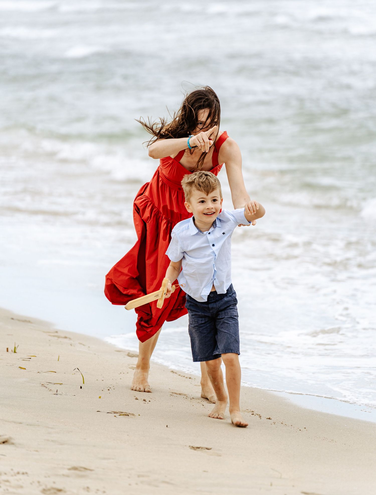 Mother in red dress running with young boy on beach.