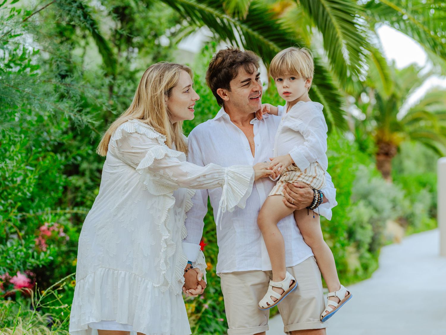 Smiling parents in white clothing hold their young child outdoors in a lush garden.