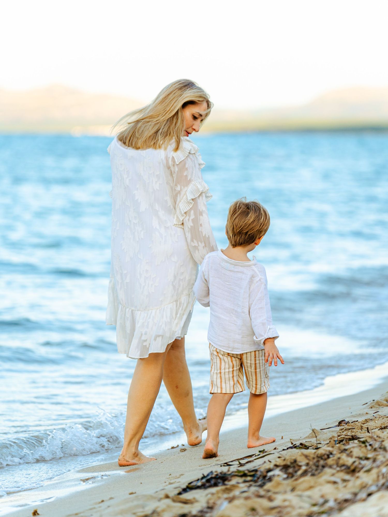Mother and young son in white outfits strolling along calm beach shoreline at golden hour.