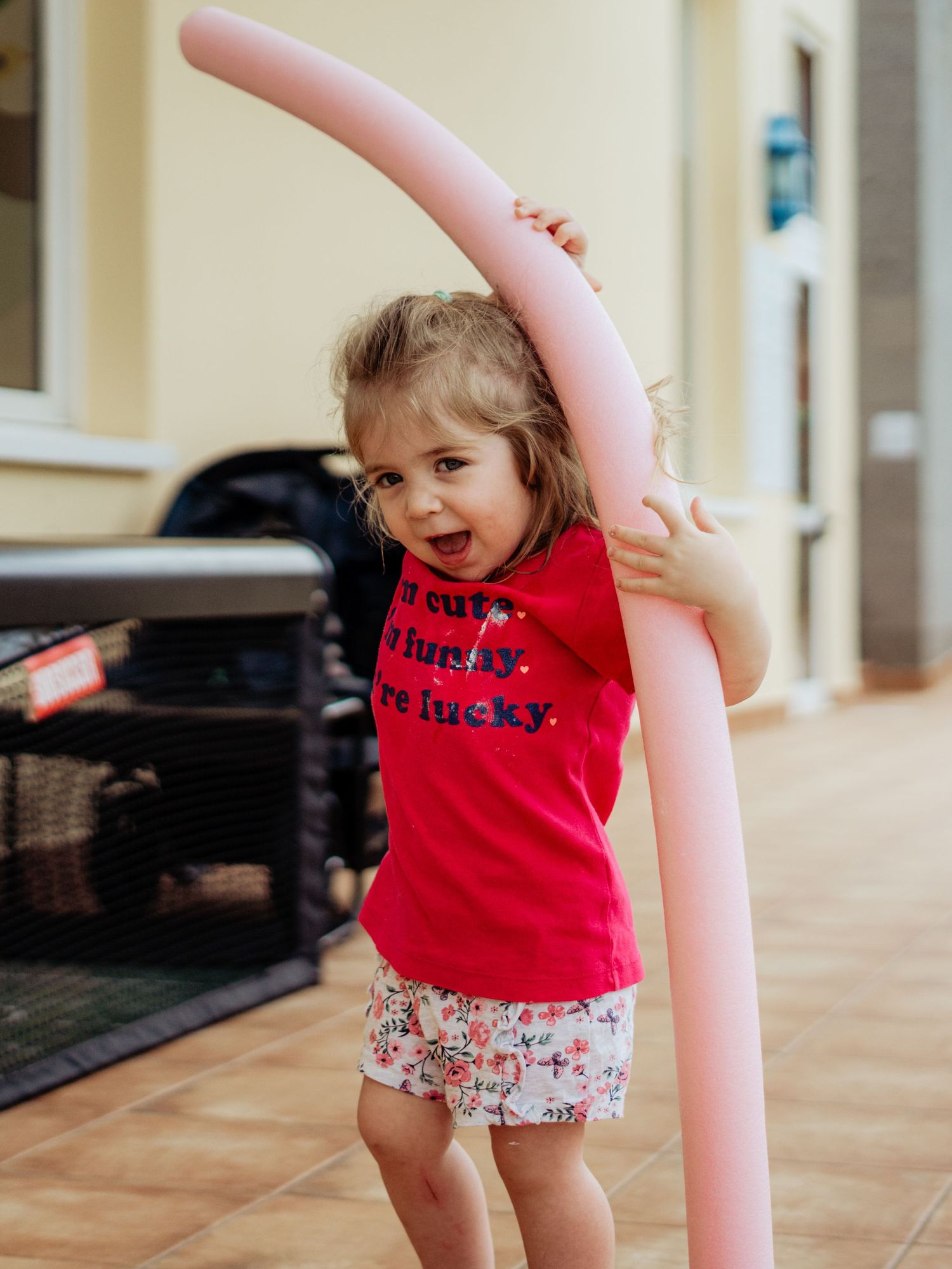 Laughing toddler girl in red top hugging a tall pink pool noodle on an outdoor patio.