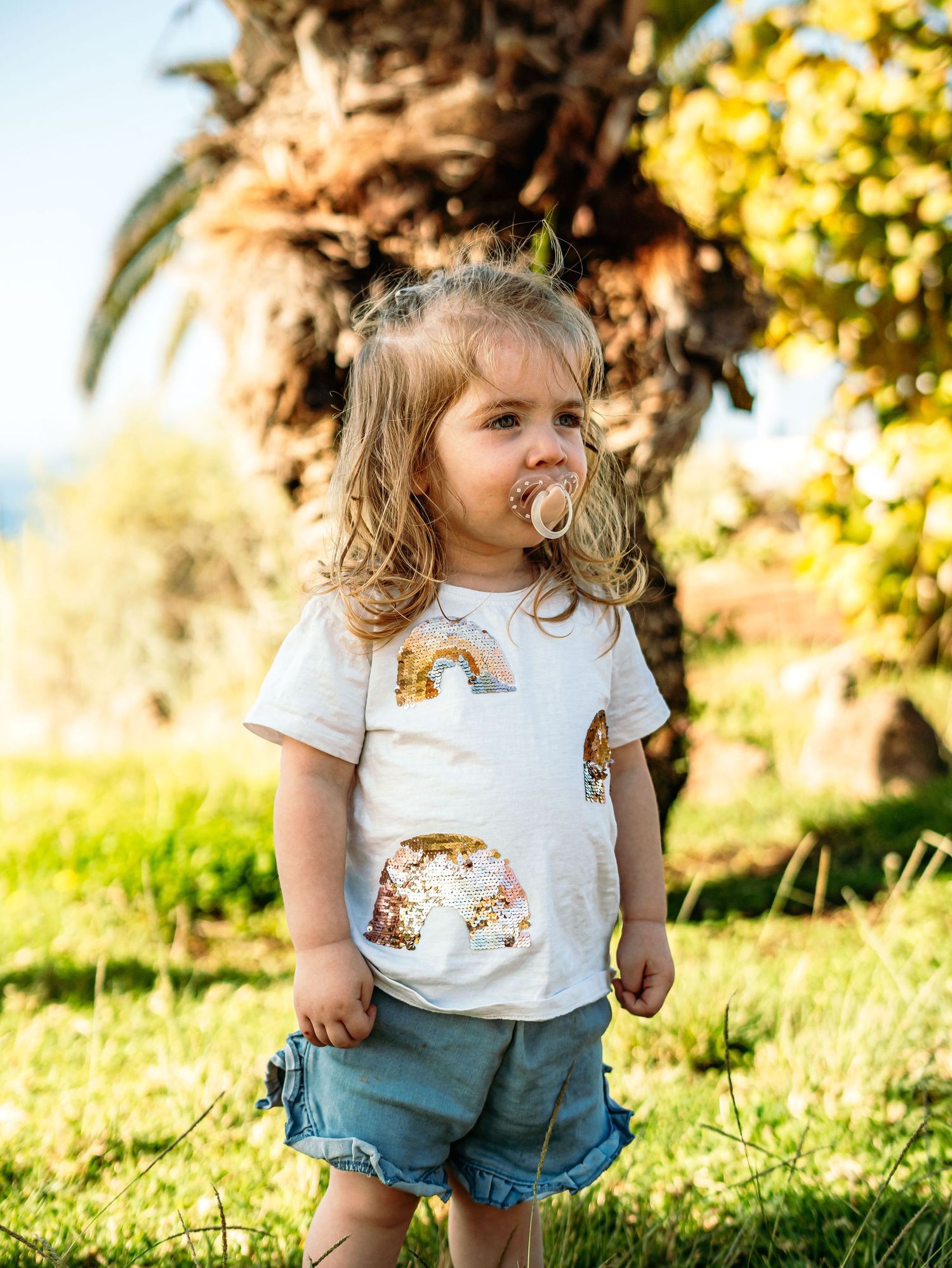 Toddler girl with pacifier in rainbow graphic t-shirt and denim shorts standing by palm tree.