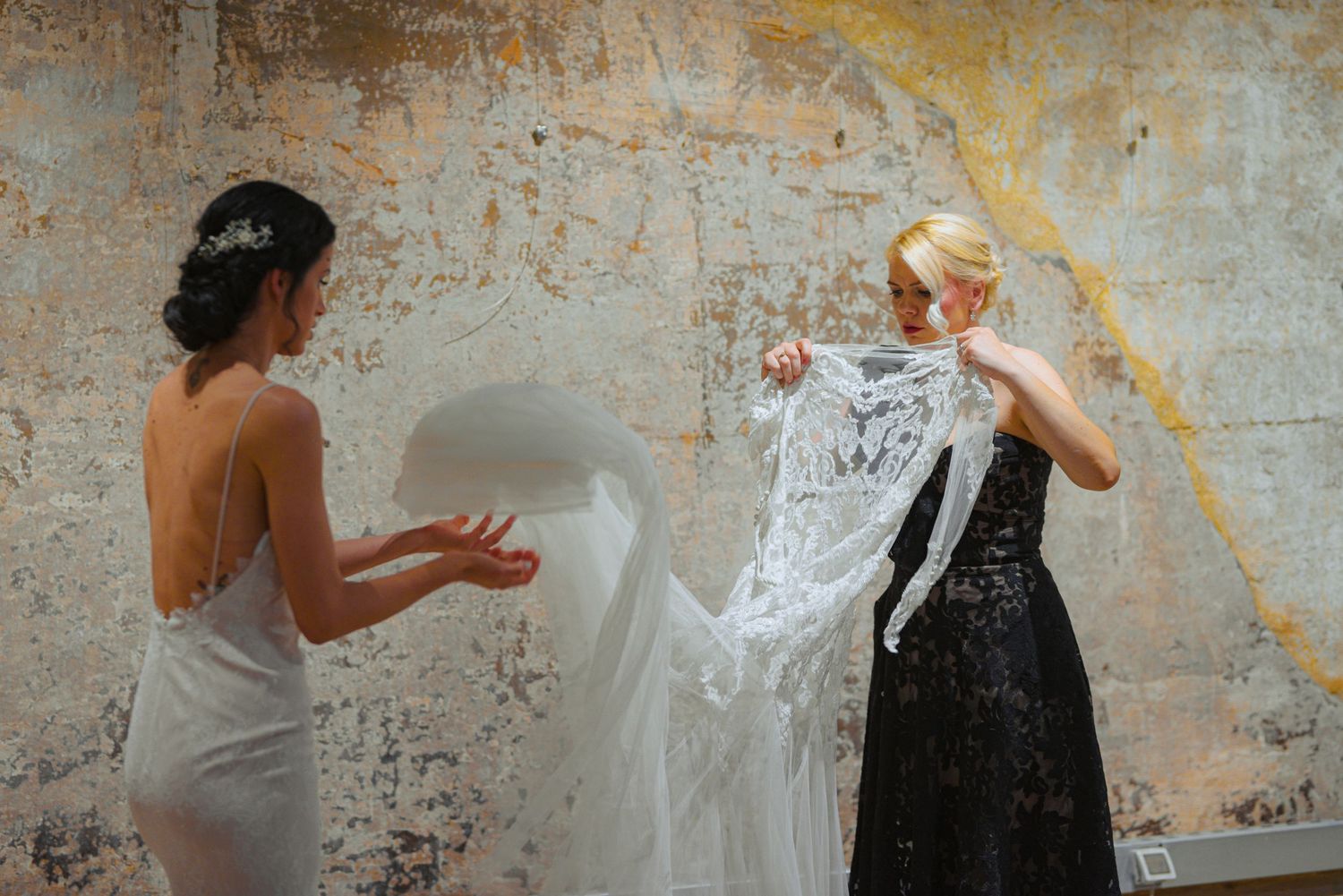 Bride and bridesmaid handling elegant lace wedding veil against rustic textured wall.