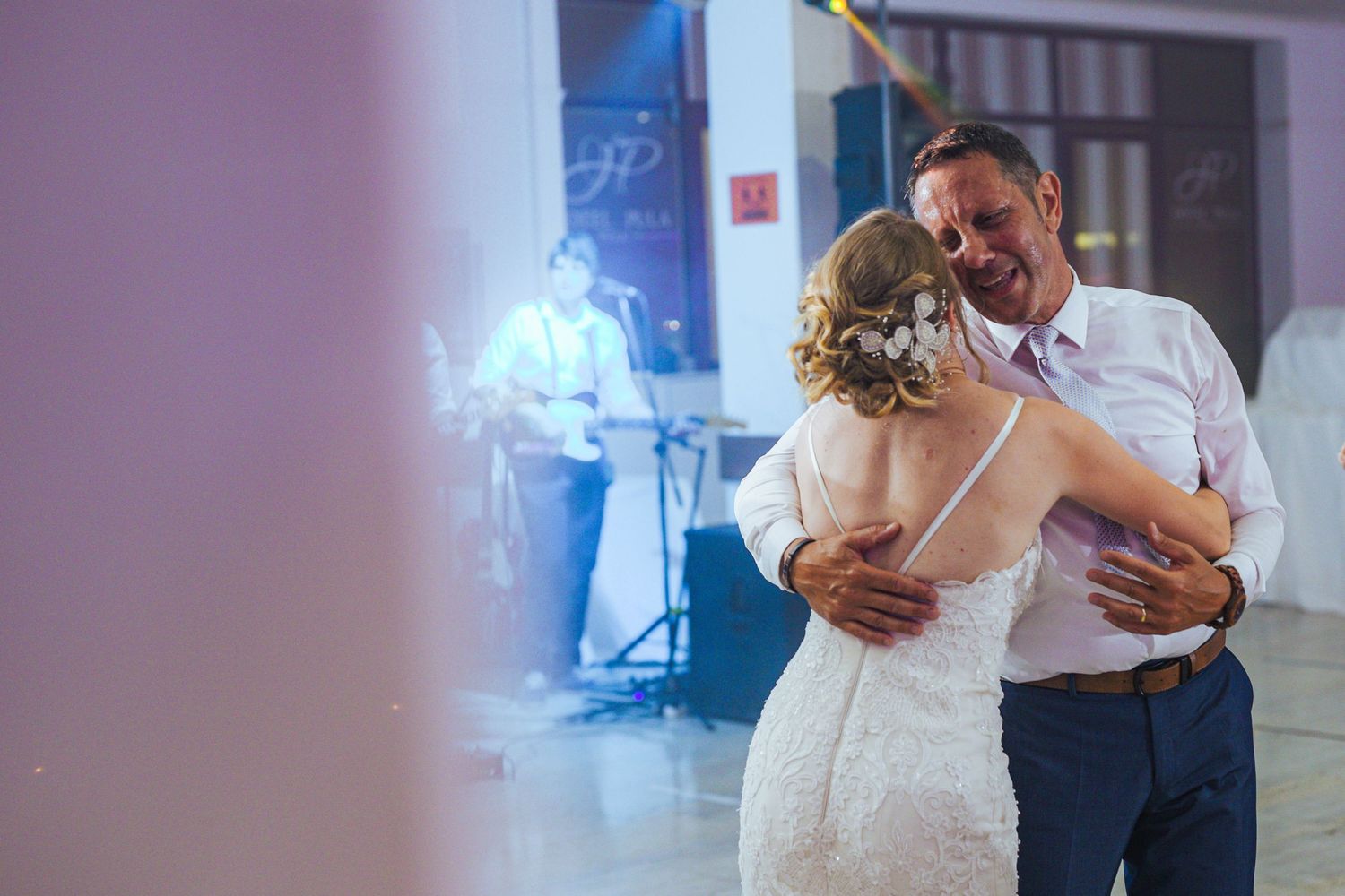Bride in lace gown shares emotional dance with older man at wedding reception with live band.