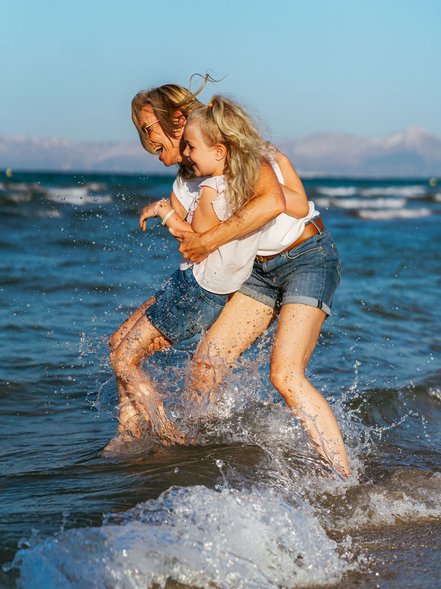 Joyful mother carrying her laughing daughter on her back while splashing through ocean waves at the beach.