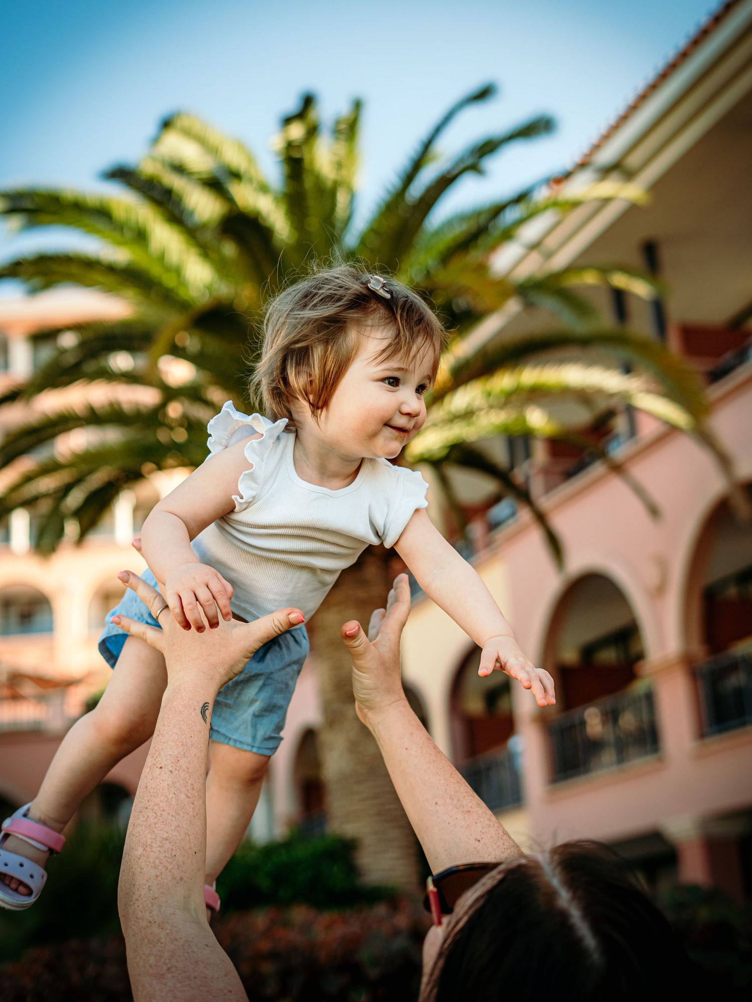 Laughing baby girl is joyfully lifted high in the air by parents with a tropical palm tree backdrop.