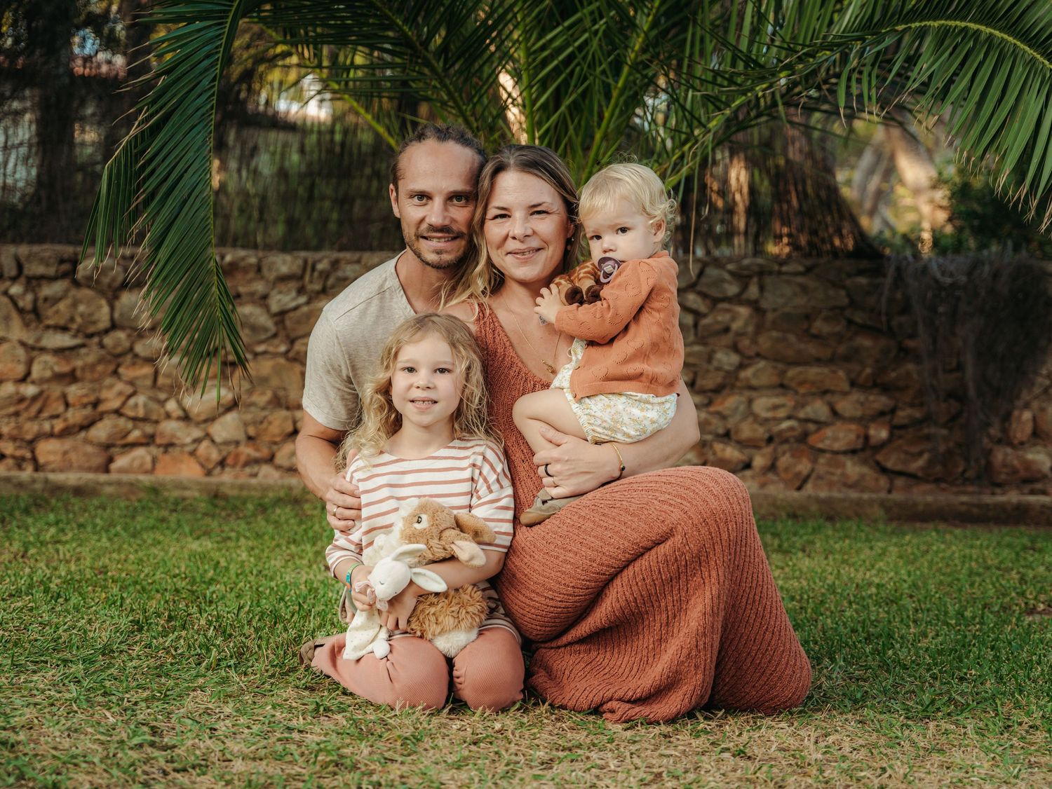 Happy family of four posing outdoors on grass near stone wall and palm tree.
