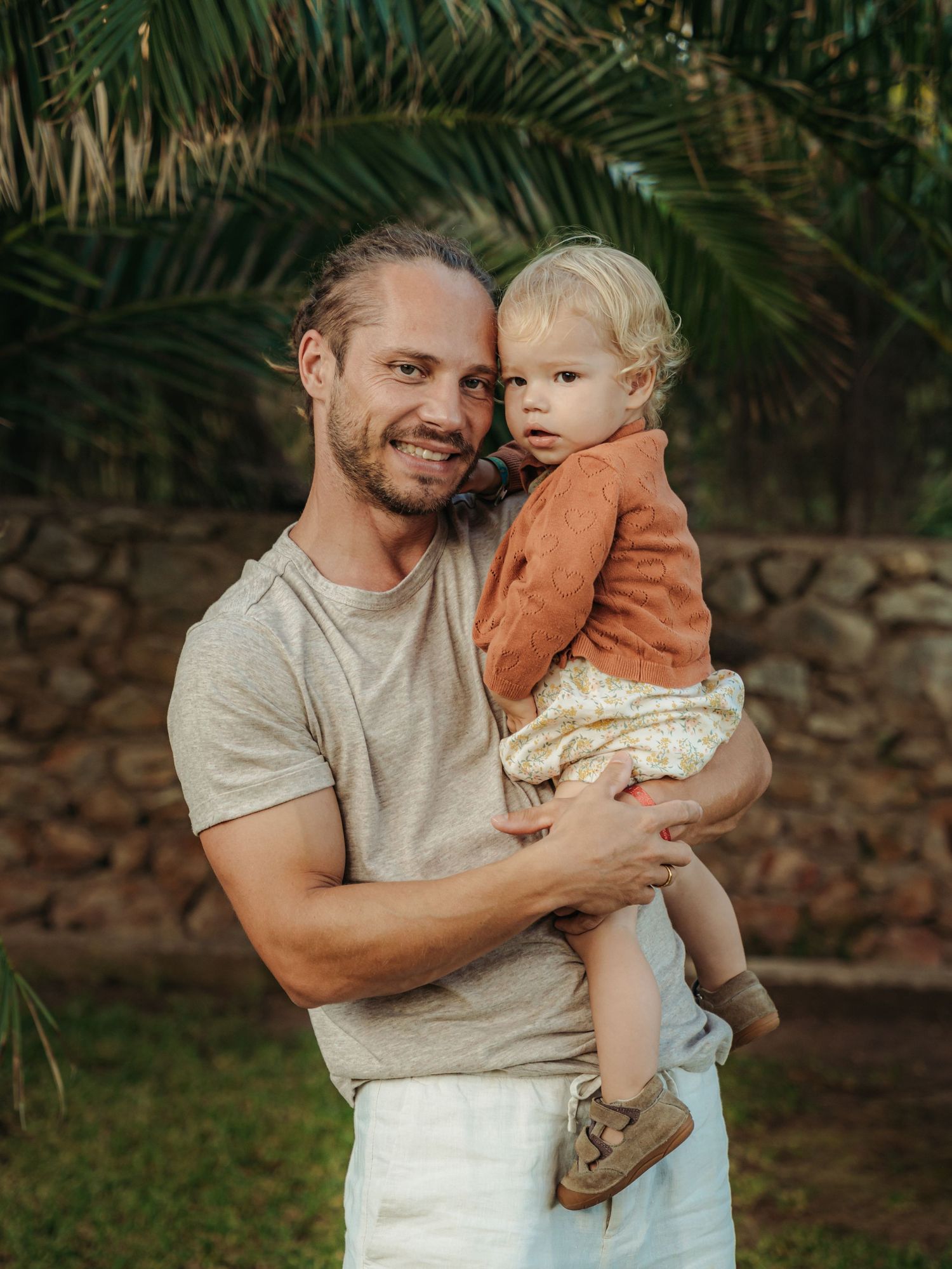 Smiling father with tied-back hair holding young blonde toddler in a garden with palm trees.