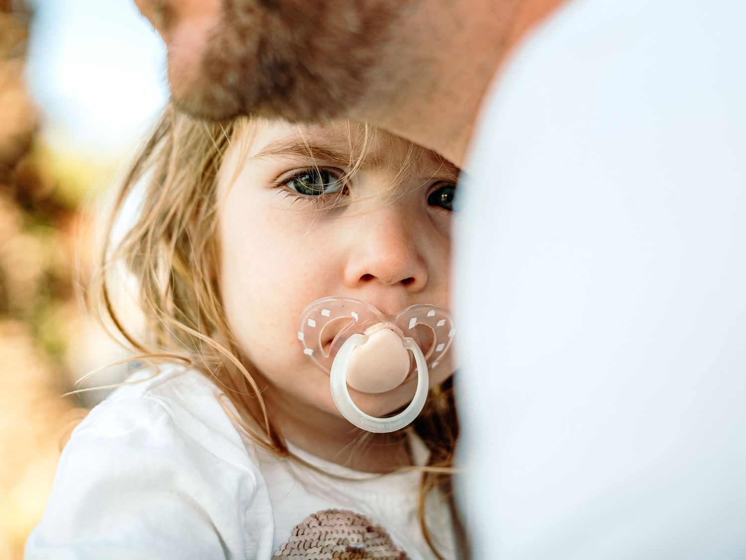 Close-up portrait of a young child with blue eyes holding a pacifier, partially hidden by an adult.