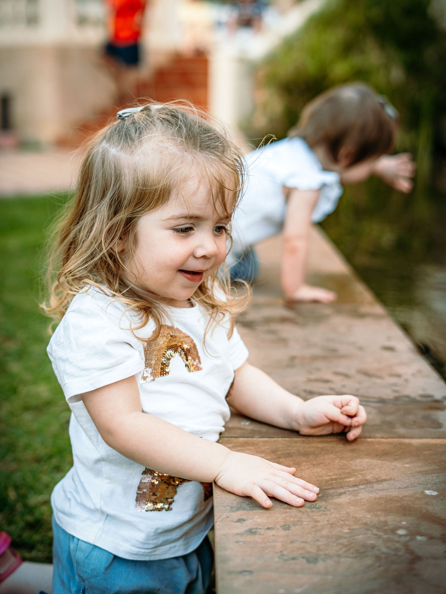 Cheerful blonde toddler girl in sequin shirt playing by a water feature with sibling.