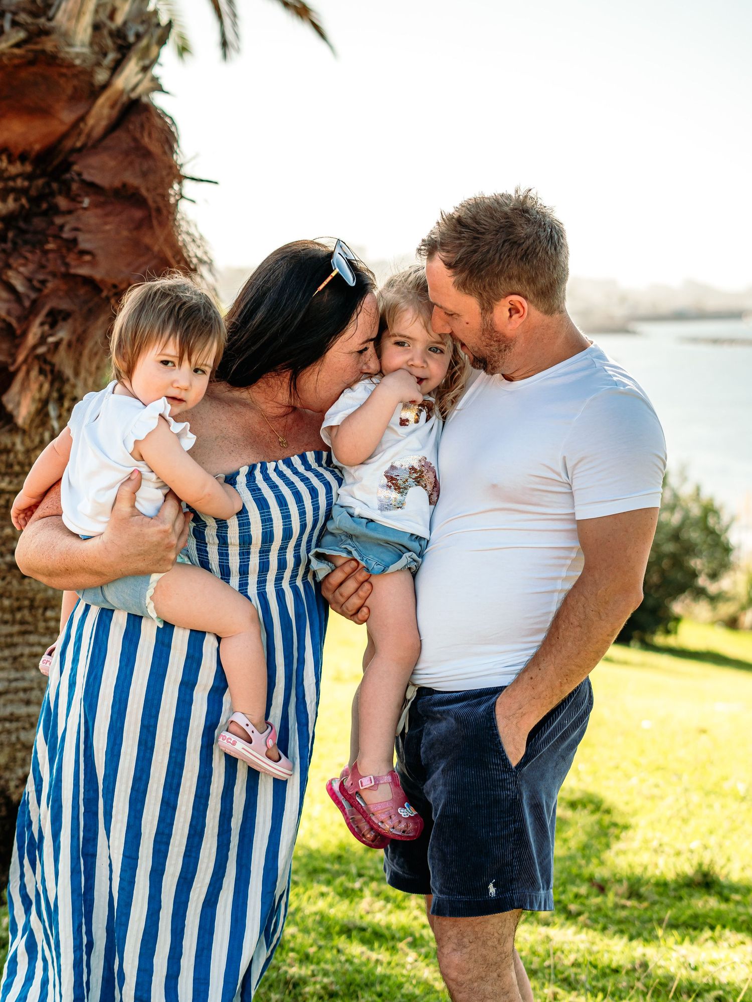 Loving family of four sharing kisses and cuddles near a palm tree by the waterfront.