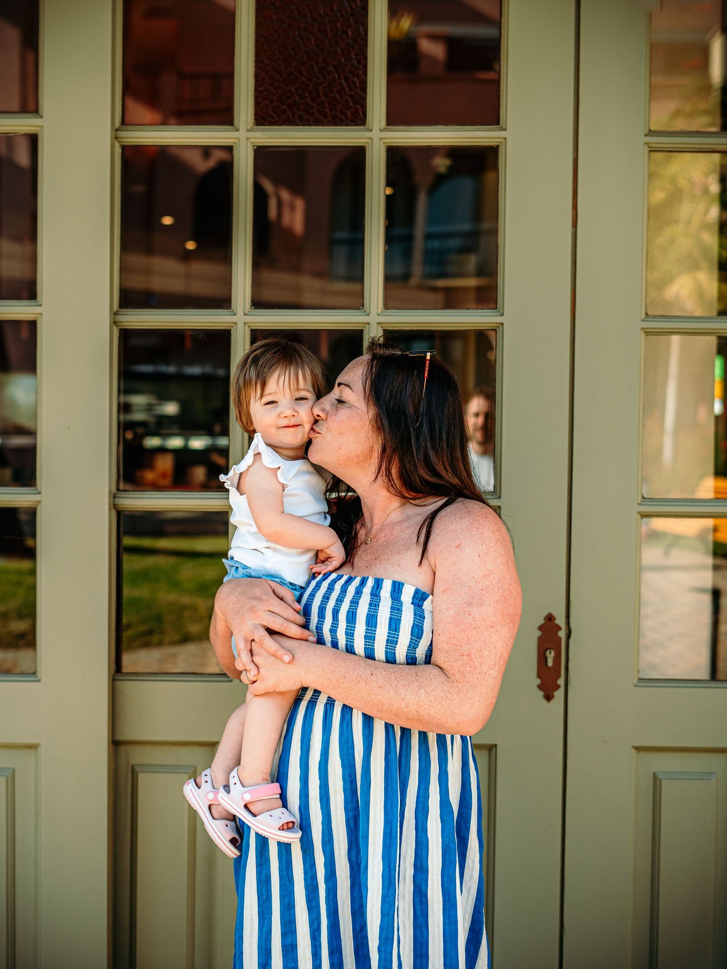 Mother in blue striped sundress kissing her baby girl on the cheek while holding her outdoors.