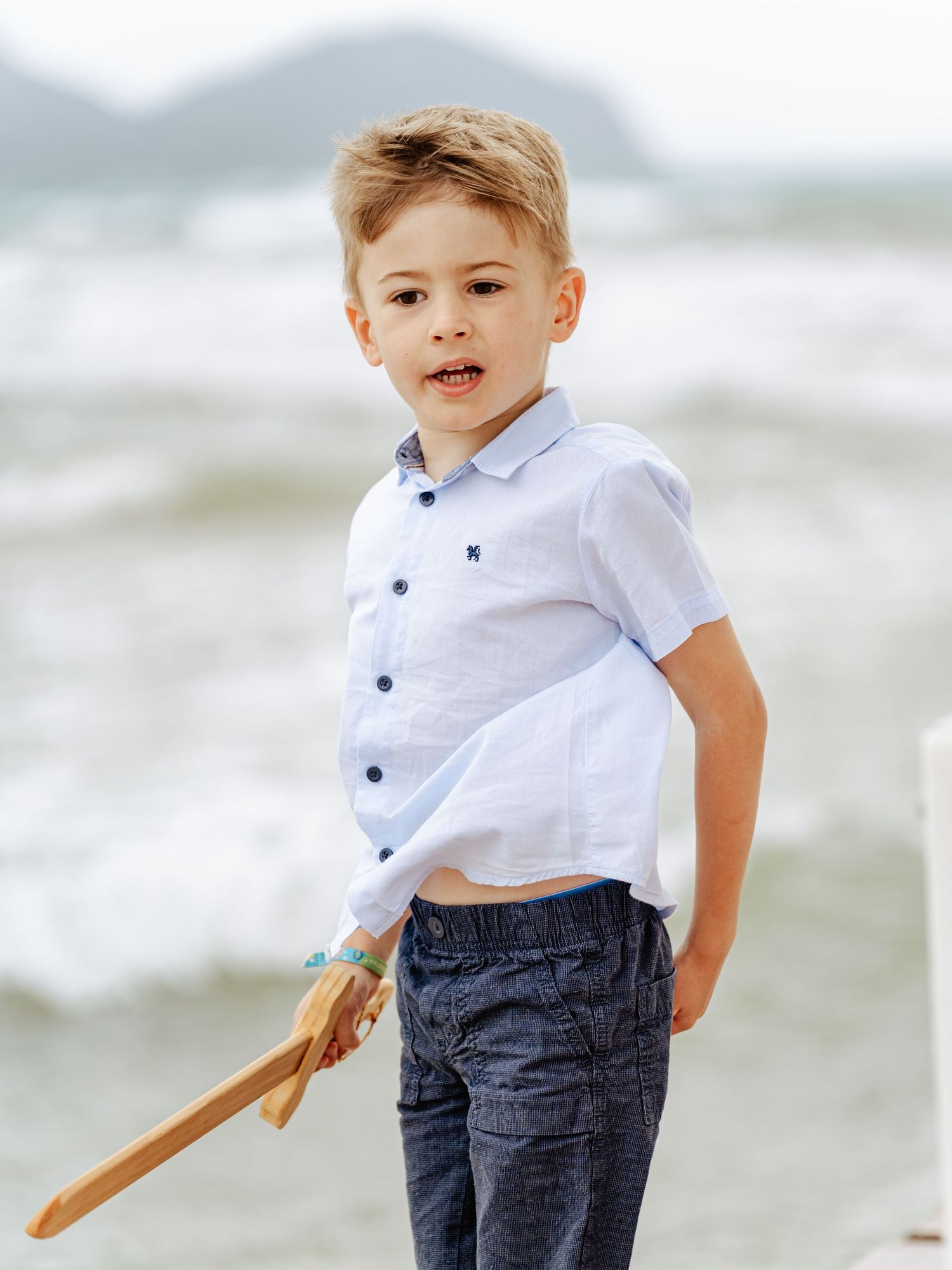 Smiling young boy in light blue shirt holding wooden toy on beach.